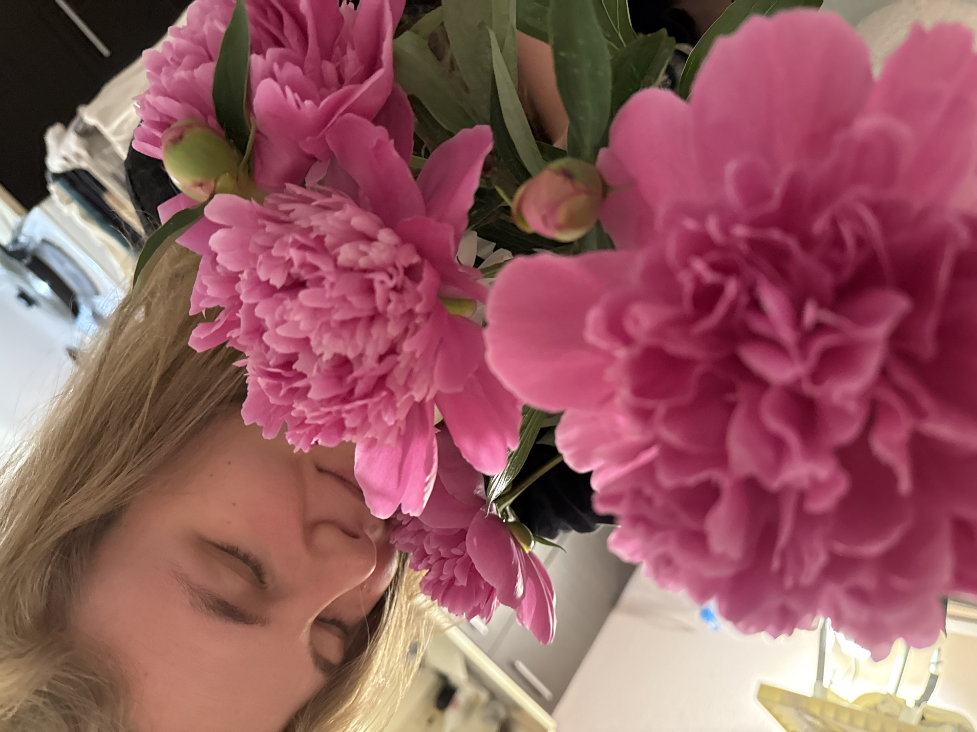 The image displays a female subject smelling a bouquet of pink peonies indoors in the city of Vladimir, Russia. The individual, with light-colored hair, is positioned on the left side of the frame. Her eyes are closed, and her face is oriented towards a cluster of vibrant, fully bloomed pink peonies, indicating an act of smelling. A large pink peony is prominent in the immediate foreground, intentionally blurred. Other pink peonies, along with at least two distinct green peony buds and green foliage, are visible in the mid-ground around the woman's face. The subject is wearing a dark-colored top. The background is out of focus, depicting an interior domestic environment with light-toned surfaces and indistinct objects, consistent with a kitchen or living area.
