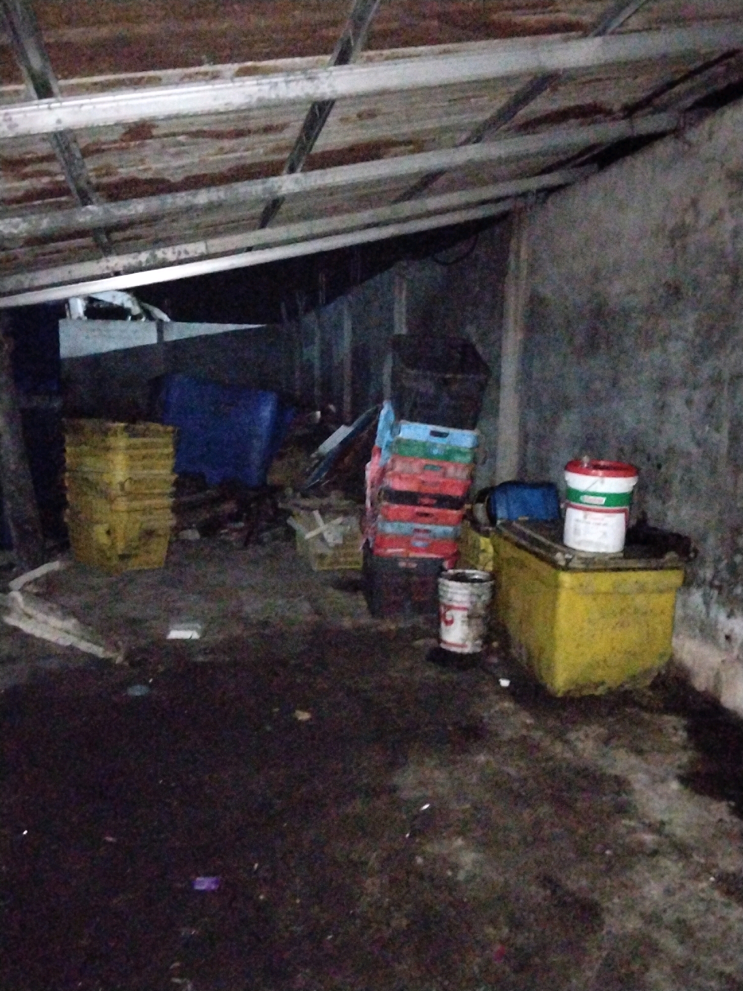 The image depicts the interior of a dimly lit storage area, possibly an abandoned or disused building, located in Moro, Indonesia. The scene is cluttered and shows signs of disrepair. The ceiling is constructed with corrugated metal roofing supported by exposed metal beams. The walls appear to be concrete.
In the foreground, the ground is a dark, uneven surface, possibly dirt or concrete, with scattered debris. To the left, a stack of yellow plastic crates is visible. Behind these crates, a large blue tarp or plastic sheeting is draped over something. Further to the right, a large yellow plastic container or tank sits on the ground. On top of this yellow container are two buckets. The top bucket is white with red and green bands, and the bottom bucket is white with red lettering. A smaller, partially visible white bucket with red markings is on the ground in front of the yellow container.
To the right of the blue tarp, a stack of colorful plastic crates (red, green, blue, and possibly white) is arranged vertically. Behind this stack, a dark, tall structure, possibly another large container or storage unit, is visible.
The lighting in the scene is poor, casting long shadows and obscuring details in some areas. The overall impression is one of disarray and neglect. There are no people visible in the image.