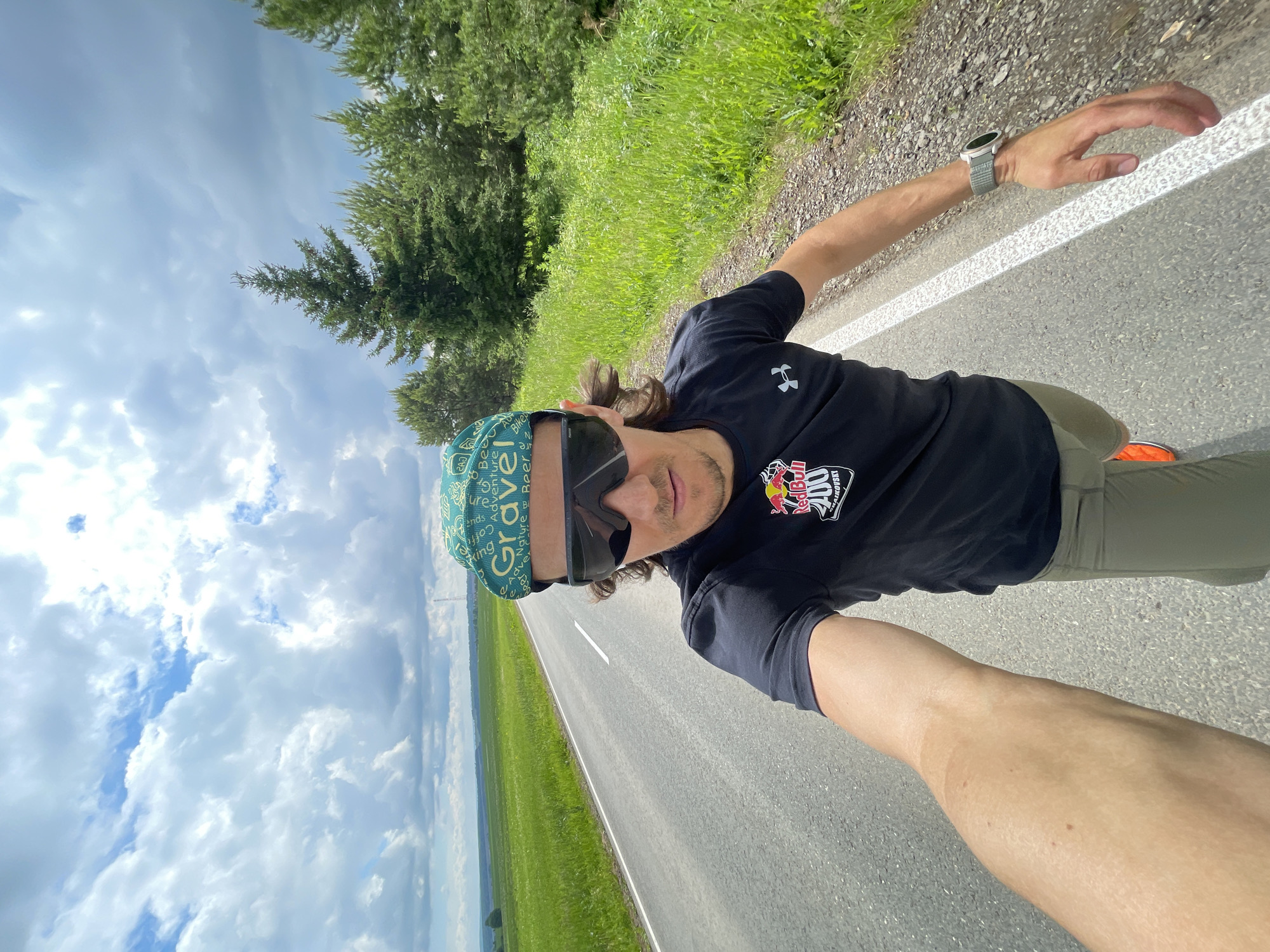 A man is captured in a dynamic selfie, with the image rotated 90 degrees clockwise. He faces the camera directly, wearing large black sunglasses, a green and teal bandana with the word "GRAVEL" prominently displayed, and a black short-sleeved t-shirt. The t-shirt features a small Under Armour logo on the left chest and a larger Red Bull logo with "100" and "LUSA DRIVE" on the right. He has a smartwatch with a light grey band on his left wrist and wears olive green or khaki shorts. A glimpse of an orange sneaker is visible near his right foot.
The background shows a paved road with a distinct white line running along its length. To the right of the road, there's a narrow gravel shoulder, followed by vibrant green grass and dense evergreen trees. To the left, expansive green fields stretch towards a distant horizon under a vast sky. The sky is largely covered with bright, fluffy white and grey clouds, indicating an overcast but bright daytime setting. The perspective suggests an outdoor activity, possibly cycling or running, on a rural or semi-rural road. The man's expression is neutral behind his sunglasses, contributing to an active and adventurous mood.