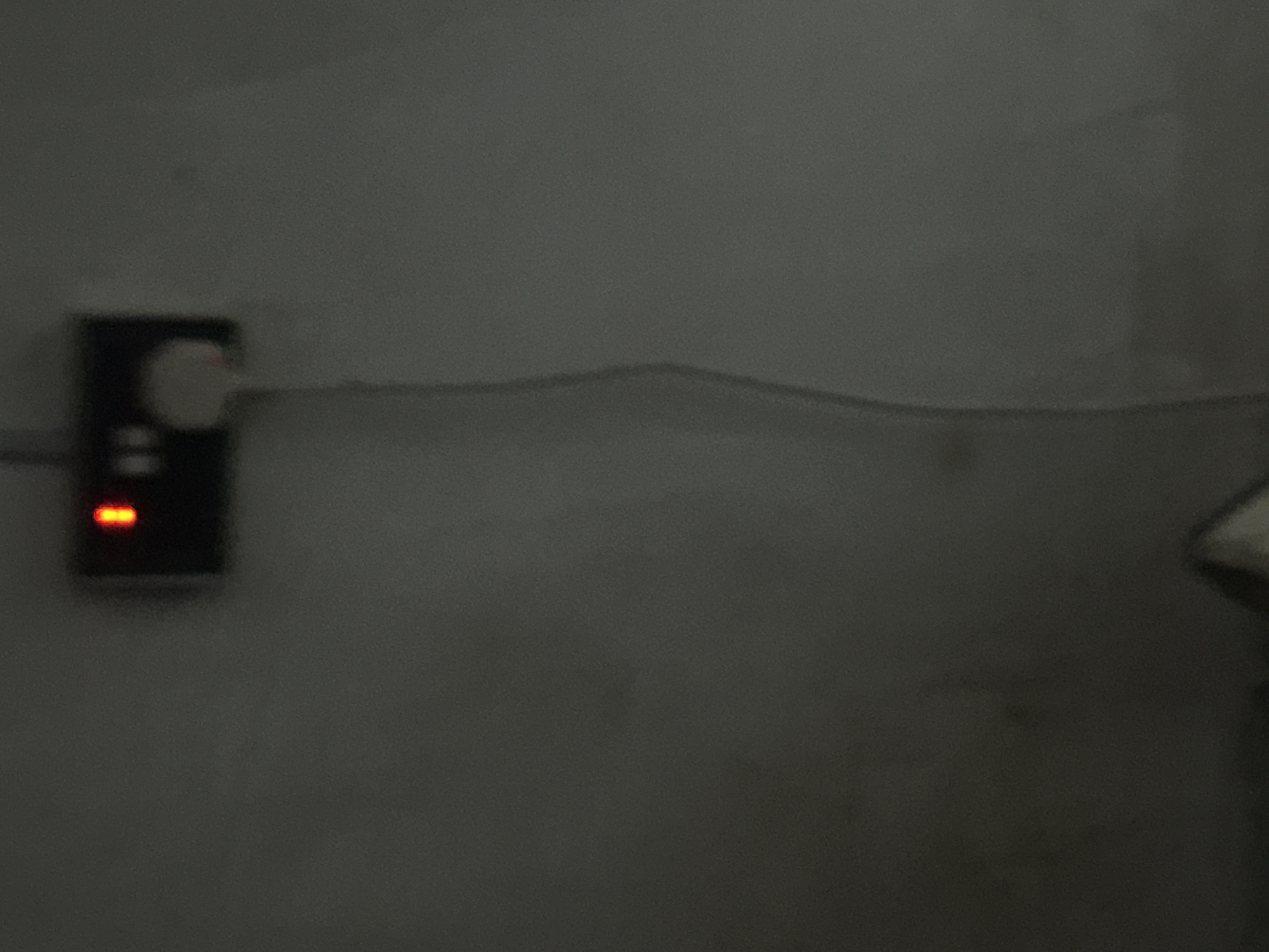 The image is a vertical, slightly blurred shot taken in low light conditions. In the upper center of the frame, a black electrical outlet with a red indicator light is mounted on a dark, textured wall. A white cord extends downwards from the outlet, curving slightly. The rest of the image consists of a gray, featureless wall. There are no people or other discernible objects in the scene. The location is described as Unknown, Unknown.