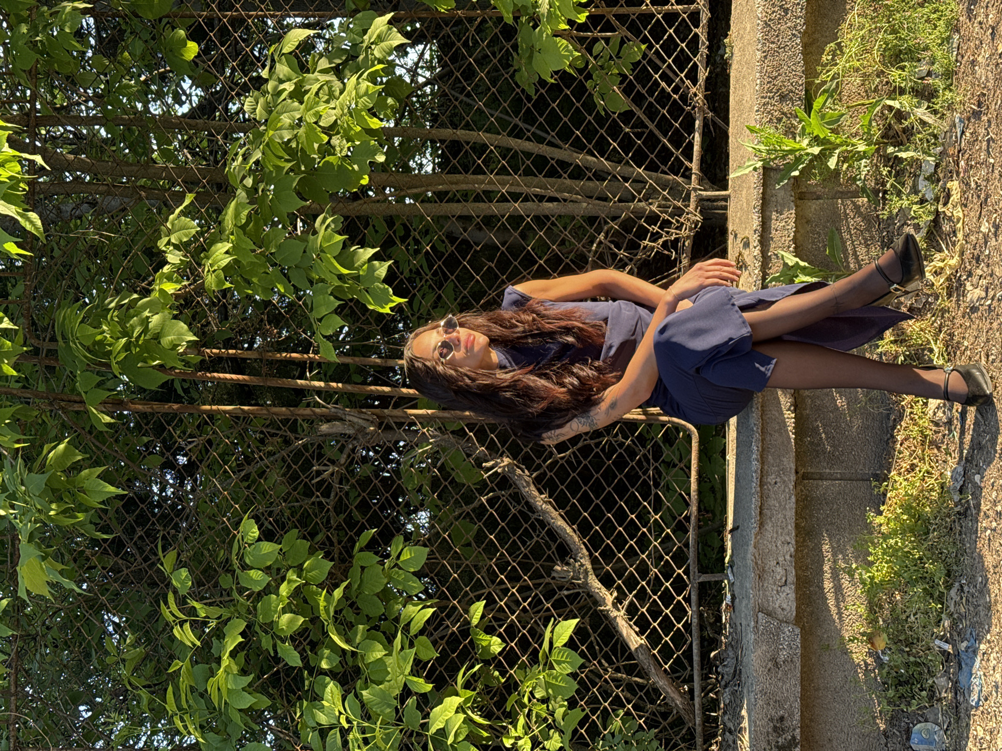 A woman with long brown hair and sunglasses is reclining on a ledge beside a chain-link fence overgrown with foliage. She is wearing a dark blue dress and black sheer stockings with black Mary Jane-style shoes. She is facing towards the viewer, but slightly angled to the right. Her arms are bent at the elbows, with her hands resting near her waist. The background consists of the chain-link fence, lush green foliage, and a concrete ledge separating it from a dirt and grassy area. The scene appears to be outdoors, likely in a park or a roadside area within the city of Pleven, Bulgaria.