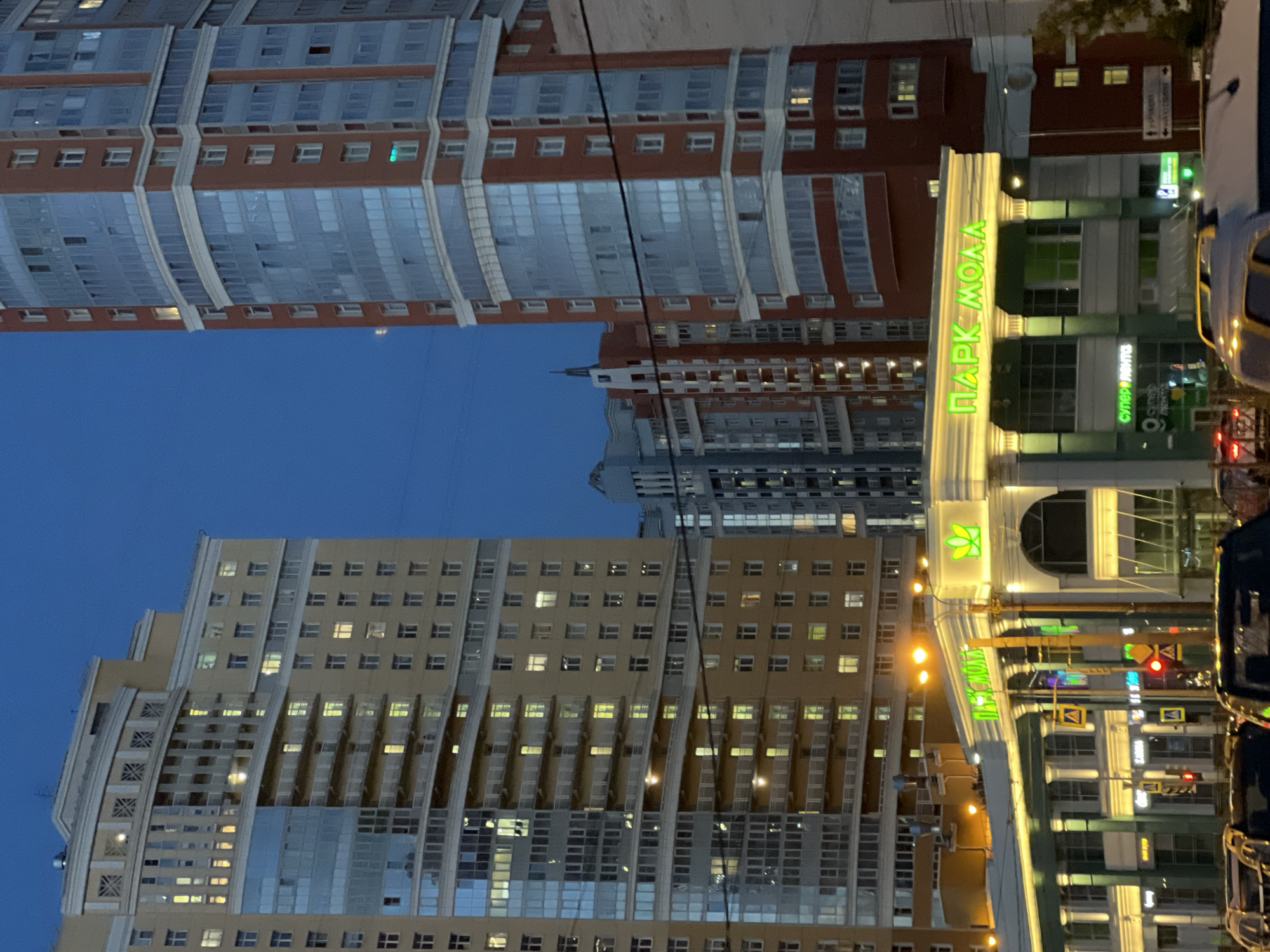 The image depicts an urban scene at dusk or night in Ladozhskoe Ozero, Russia. Multiple modern high-rise buildings dominate the frame, their facades illuminated by artificial lights. The buildings are a mix of residential and commercial structures, characterized by their repetitive window patterns and curved architectural elements.
In the foreground, a prominent building features a glowing green sign that reads "ПАРК МОЛЛ" (PARK MALL), indicating a shopping center. Another sign with green lettering, partially obscured, appears to say "СУПЕРМАРКЕТ" (SUPERMARKET). Traffic lights are visible, with a red light illuminated, suggesting the presence of a busy street or intersection. The overall atmosphere is one of a developed urban environment at the end of the day. The sky is a deep blue, indicating twilight or nighttime. No people are clearly visible in the image. The focus is on the architecture and commercial signage of the city.