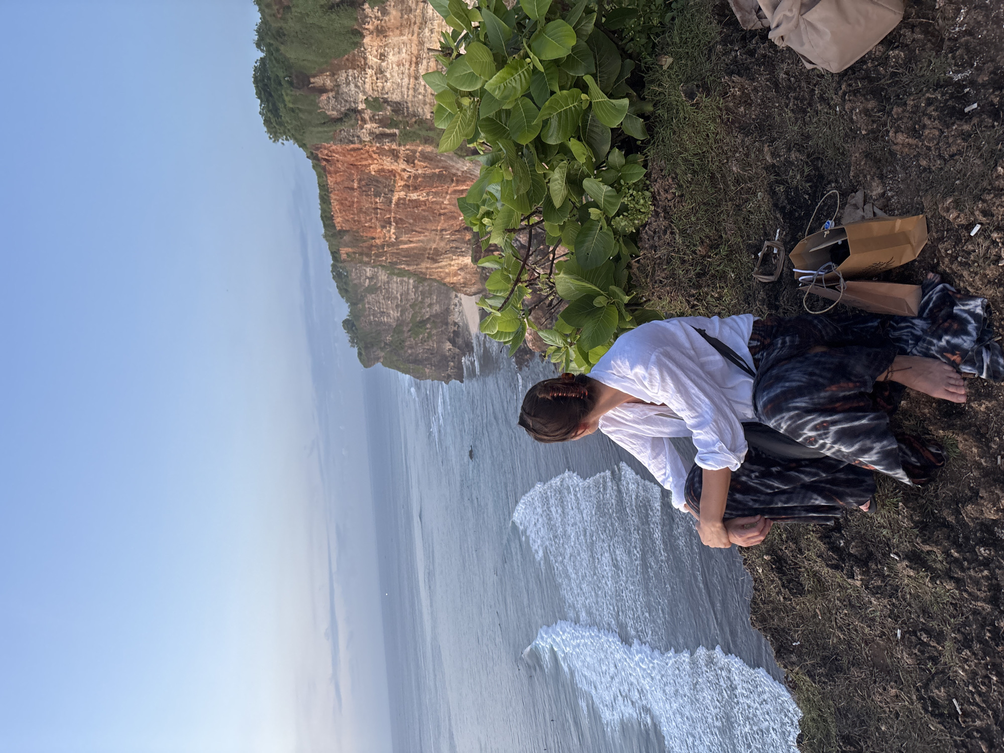 A high-angle, outdoor photograph depicts a cliff overlooking the ocean. A person wearing a white shirt and dark patterned pants sits on the edge of the cliff, looking down. To the right of the person, there are two paper bags, a tan tote bag, and some small items scattered on the ground. The cliff is covered with green vegetation. Below the cliff, waves crash against the shore. In the background, another cliff formation can be seen. The sky is light blue and partially visible. The location is identified as Kuta Selatan, Indonesia.