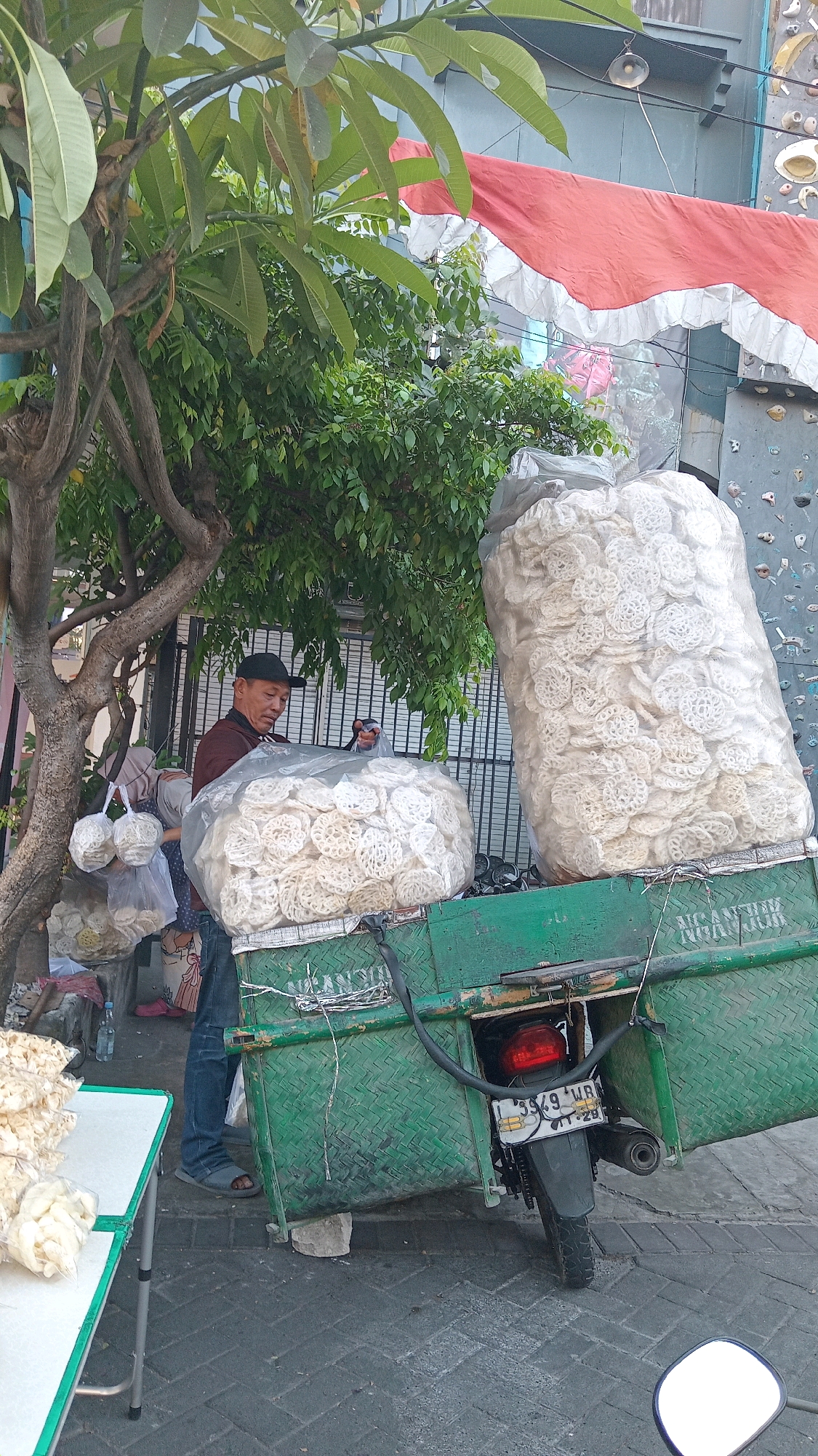 An outdoor scene in the city of Randuagung, Indonesia, features a man in a maroon shirt and blue jeans, wearing a black baseball cap, standing next to a green motorcycle with a trailer. The trailer is laden with large, clear plastic bags filled with white, circular crackers. One bag is particularly massive, overflowing onto the trailer. To the left of the man, a table with a white top and green trim displays smaller bags of crackers. A tree with green foliage shades a portion of the scene. In the background, a building with a wall that appears to be a climbing wall and a red and white banner are visible. The ground is paved with gray bricks. A license plate on the motorcycle reads "L 3949 WR". The man is holding a plastic bag, possibly packaging crackers. Another person, partially obscured, stands behind him, also appearing to be involved with the cracker sale. A water bottle and more bags of crackers are on the ground near the table.