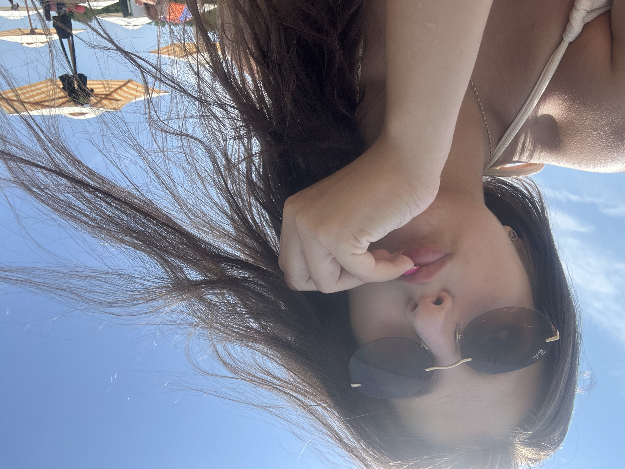 A young woman is taking a selfie outdoors on a sunny day. She is wearing a bikini top and sunglasses. Her hair is long and dark and appears to be blowing in the wind. Her lips are slightly parted and she has her finger resting on them. In the background, there are beach umbrellas and people. The location is the beach in Varna, Bulgaria.