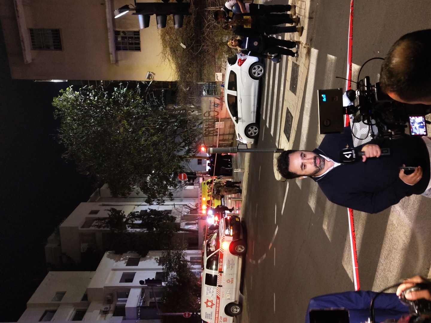The image depicts a nighttime urban scene in Tel-Aviv, Israel, centering on an apparent incident or news event.
In the immediate foreground, a bearded male reporter, wearing a dark blue long-sleeved shirt, stands facing slightly left, holding a black microphone with a "K1" logo and Hebrew text. He appears to be reporting, with a professional video camera on a tripod to his right, operated by an individual whose head is visible in the lower right corner. A red and white barrier tape runs horizontally across the lower third of the frame, separating the foreground from the street.
The midground features a street with various activities. On the far left, a white Magen David Adom ambulance with "TEL AVIV" and "MAGEN DAVID ADOM" in Hebrew (numbers 101, 1930) is parked with its red and blue emergency lights flashing. Behind it, another emergency vehicle with yellow flashing lights is partially visible. Individuals in what appear to be military or security uniforms are seen near these vehicles. On the right side of the street, a white hatchback car (resembling a Chevrolet Spark) is stationary on a pedestrian crosswalk. A group of four to five individuals stands on the sidewalk near the white car; at least one woman wears a dark blue uniform with reflective strips, and another individual holds a helmet. Road markings, including dashed lines and a crosswalk, are visible on the asphalt. A traffic light with red and amber lights is on the far right, and a red circular "no entry" sign is visible further back.
The background reveals multi-story buildings, predominantly light-colored, with numerous windows. A large, dense-foliaged tree dominates the center-right background, partially obscuring a building. Streetlights and ambient illumination from the buildings light the scene, under a dark night sky.