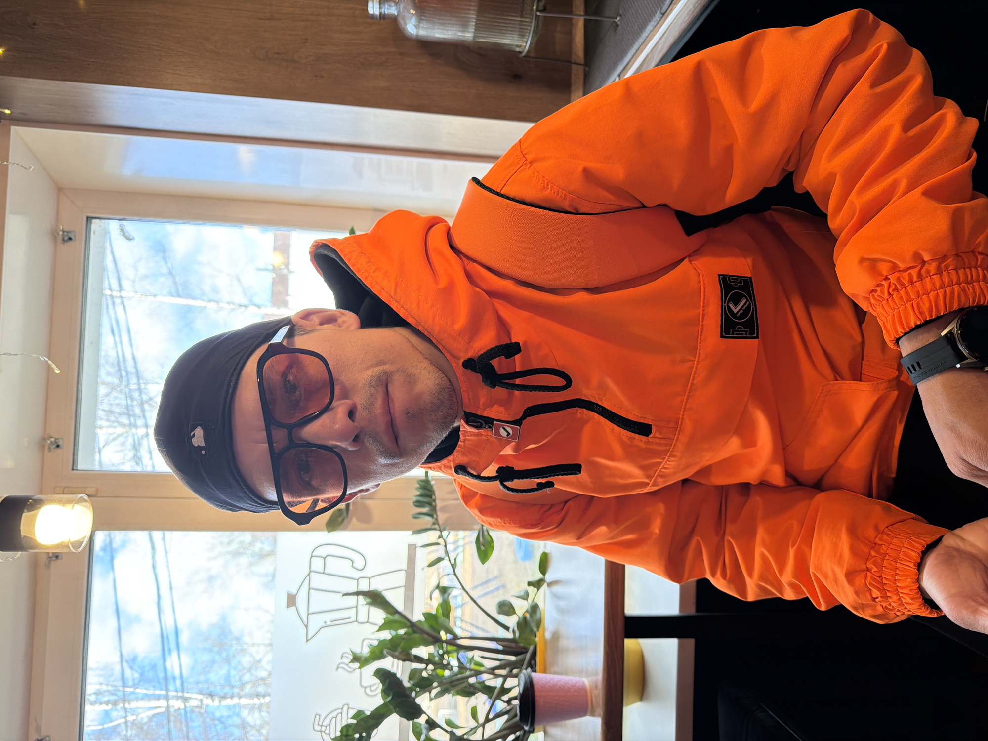 The image captures a man indoors, looking directly at the viewer. He wears a vibrant orange hooded jacket, a black beanie, and black-framed glasses with orange-tinted lenses. He has short facial hair and a dark-colored smartwatch on his left wrist.
The man is positioned in front of a large window, which reveals a view of bare trees against a bright, slightly cloudy sky, indicative of winter or early spring weather during daylight hours. A white outline sticker, depicting a Moka pot (coffee maker) and possibly other coffee-themed designs, is visible on the windowpane.
Inside, to the man's right, a houseplant with green leaves sits in a pink pot on a wooden surface. A small, warm-toned light fixture is affixed to the window frame on the left. Above, part of a wooden shelf holds a ribbed glass bottle.
Notable details on the man's jacket include a black square patch on the left chest with a white, stylized symbol resembling a checkmark or a 'V' shape. A small tag attached to the main zipper pull also features a white arrow pointing right within a small square.
The overall setting suggests a casual and comfortable indoor environment, likely a cafe or a home in Chaikovsky, Russia. The man's expression is neutral. The image is oriented at a slight angle.