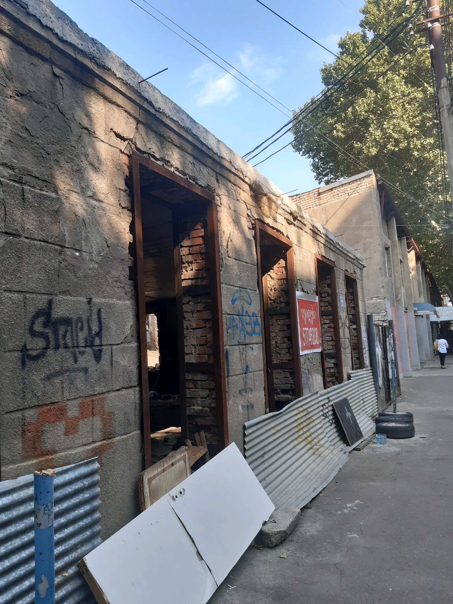 The image shows a street scene in Tbilisi, Georgia. In the foreground, there is a dilapidated building with exposed brickwork and graffiti. Several open doorways lead into dark interiors. In front of the building, there is corrugated metal sheeting, construction debris including a white door, and two tires. A person in a white shirt and dark pants is walking away from the camera down the sidewalk on the right side of the image, next to a line of columns. The sky is blue with some clouds, and a large tree with green foliage is visible in the background, along with power lines and utility poles. The overall impression is one of urban decay and ongoing construction or renovation.