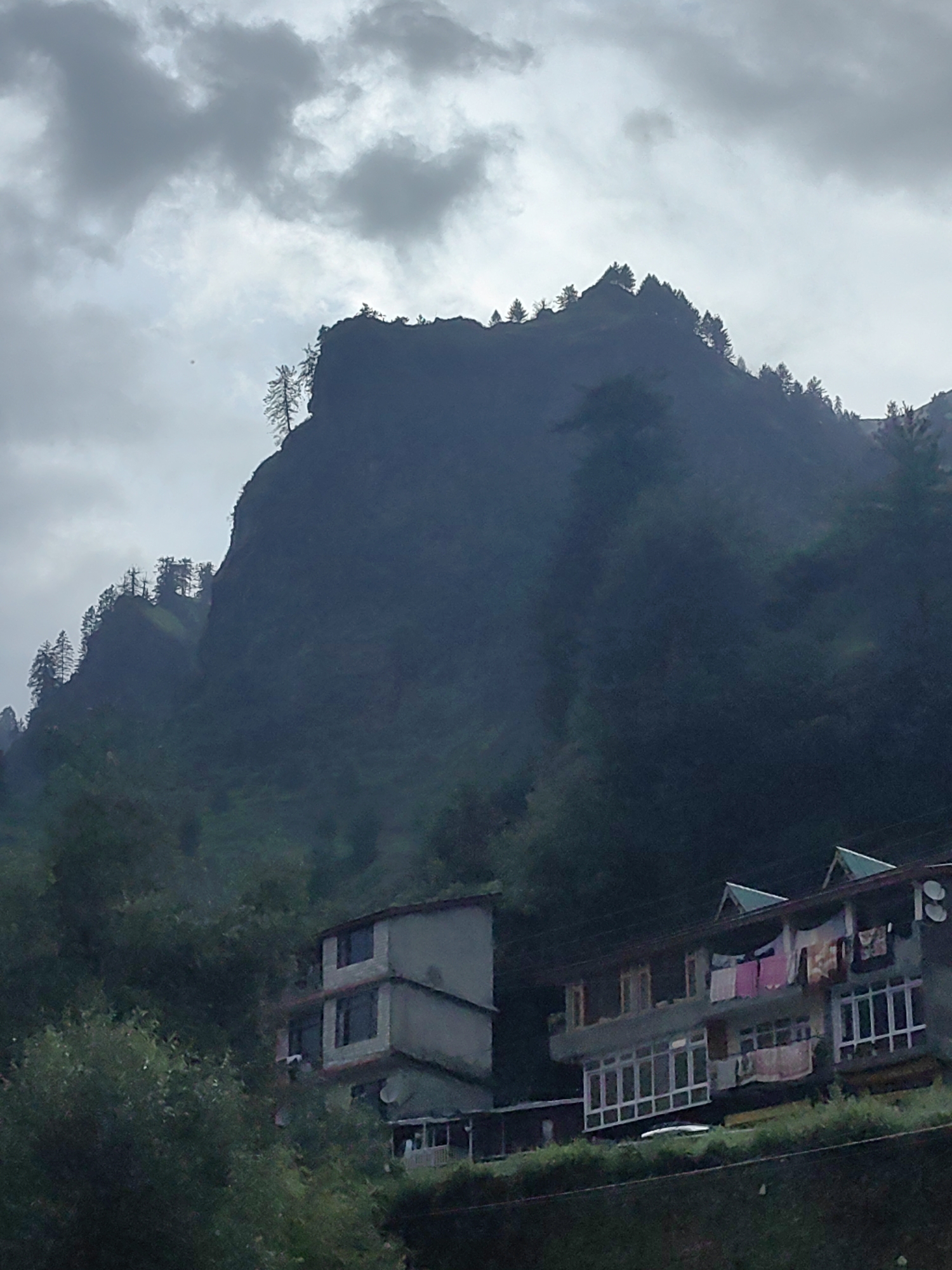 The image depicts a mountainous landscape in the city of Kullu, India. The sky is overcast with dark, cloudy conditions. In the foreground, two buildings are visible, situated on a hillside. The building on the left is a multi-story structure with multiple windows. To the right, another building features a more extended facade with numerous windows, some of which are covered by a conservatory-like structure. Laundry is hanging from the balconies of this building. The surrounding environment is characterized by lush green vegetation and trees, indicative of a natural, hilly terrain. The overall impression is one of a rural settlement nestled within a mountainous region under a cloudy sky.