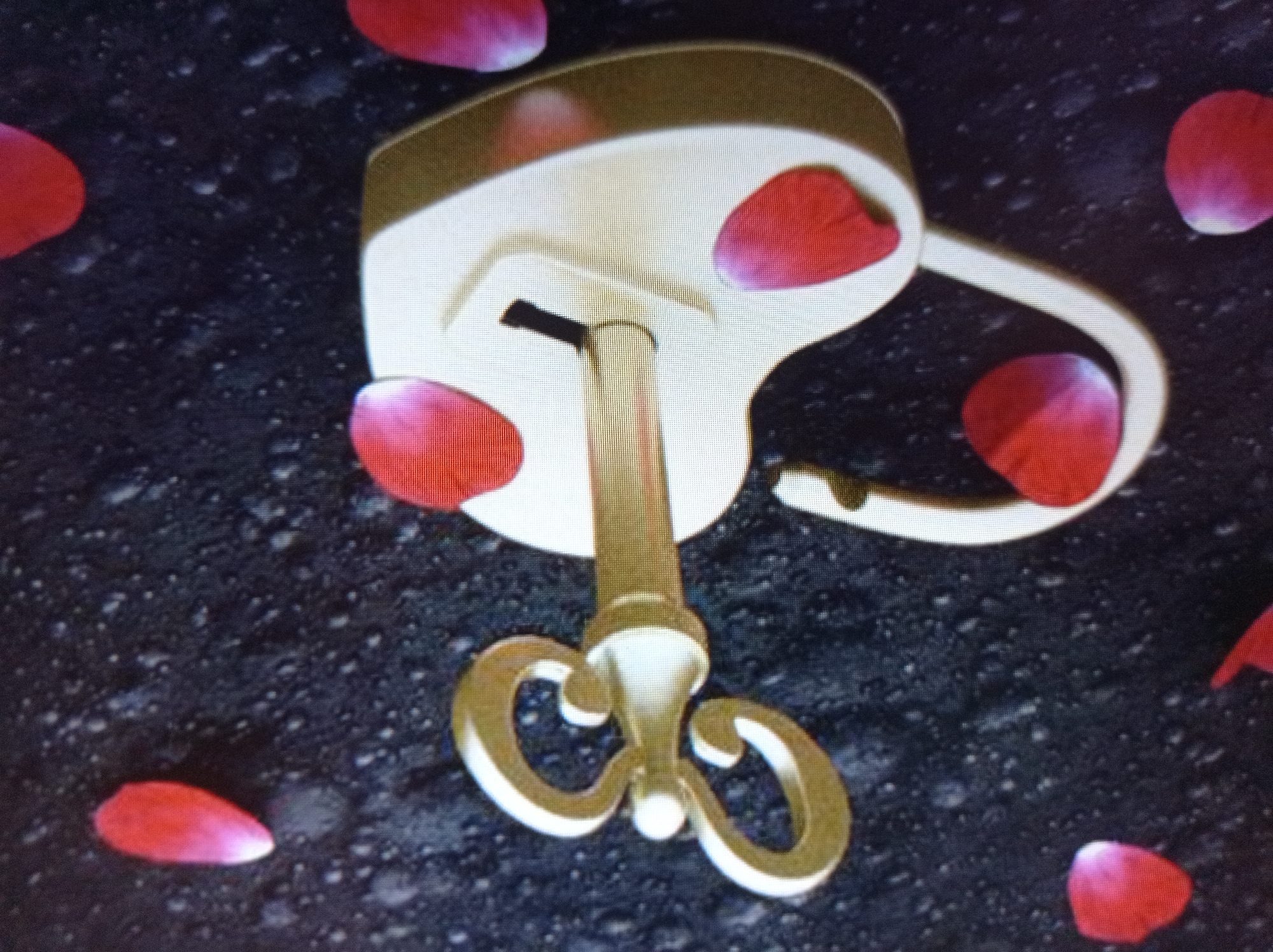 The image displays a close-up view of a golden heart-shaped padlock and key, set against a dark, textured background resembling asphalt or dark soil. Scattered around the padlock and key are several vibrant red rose petals, some appearing as if they have fallen recently. The padlock itself has a keyhole, and the key is inserted into it, suggesting a locked state. The lighting highlights the metallic sheen of the lock and key, creating reflections and depth. The overall composition evokes a romantic or symbolic theme. There is no visible indication of the city of Mukachevo, Ukraine, within the image.