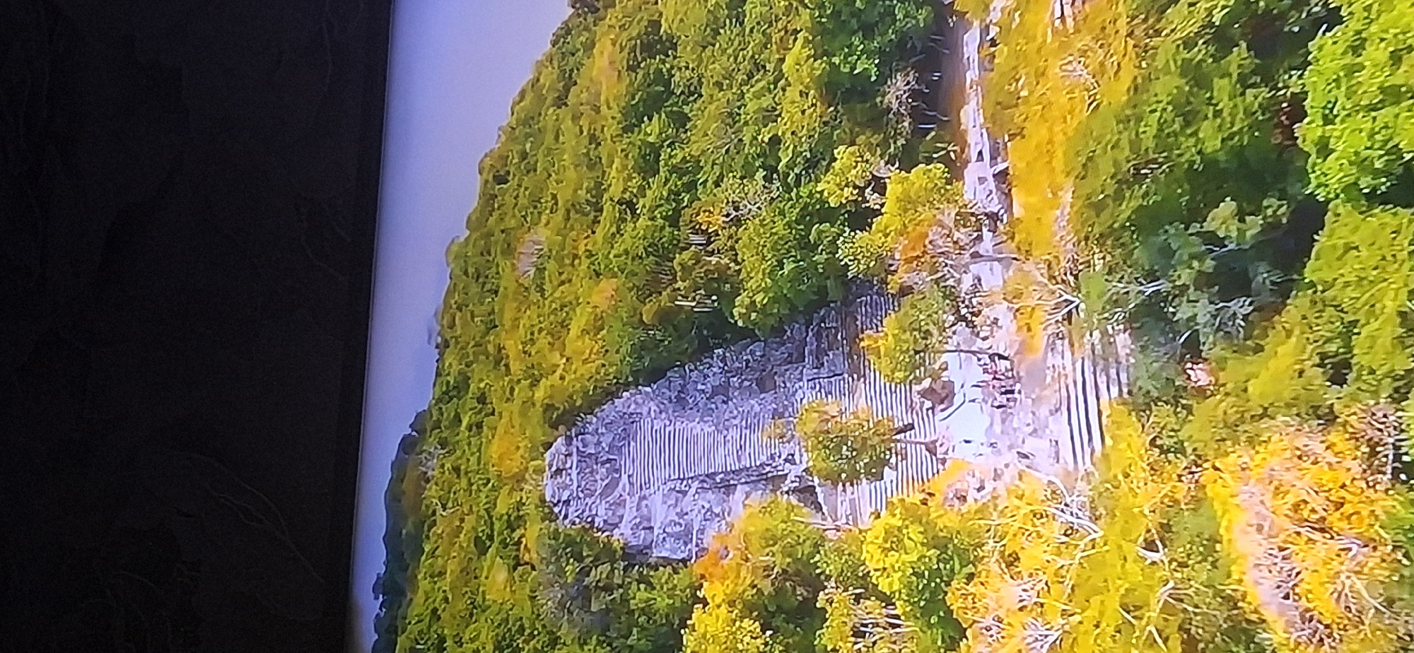 The image displays an aerial view of a stone pyramid structure with a long, wide staircase leading up its side. The structure is set within a lush, green, and yellow-tinged forest on a hillside. Several small figures, appearing to be people, are visible near the base of the staircase. The sky above is a light blue. The scene does not depict any elements related to the city of Bila Tserkva, Ukraine.