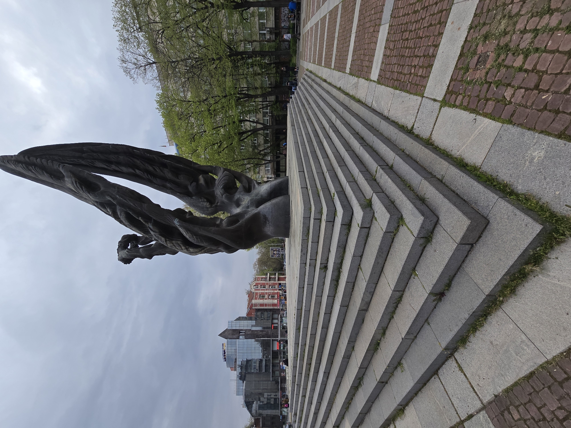 Here is a description of the image:
The image shows a bronze statue in front of a large, gray stone staircase. The statue is of a person with arms raised. Beyond the staircase, there are buildings with glass facades and red brick architecture. In the background, there are green trees and an overcast sky. The ground is paved with red brick, and there is grass growing in between the steps of the staircase. The scene appears to be in an urban area, but the exact location is unknown.