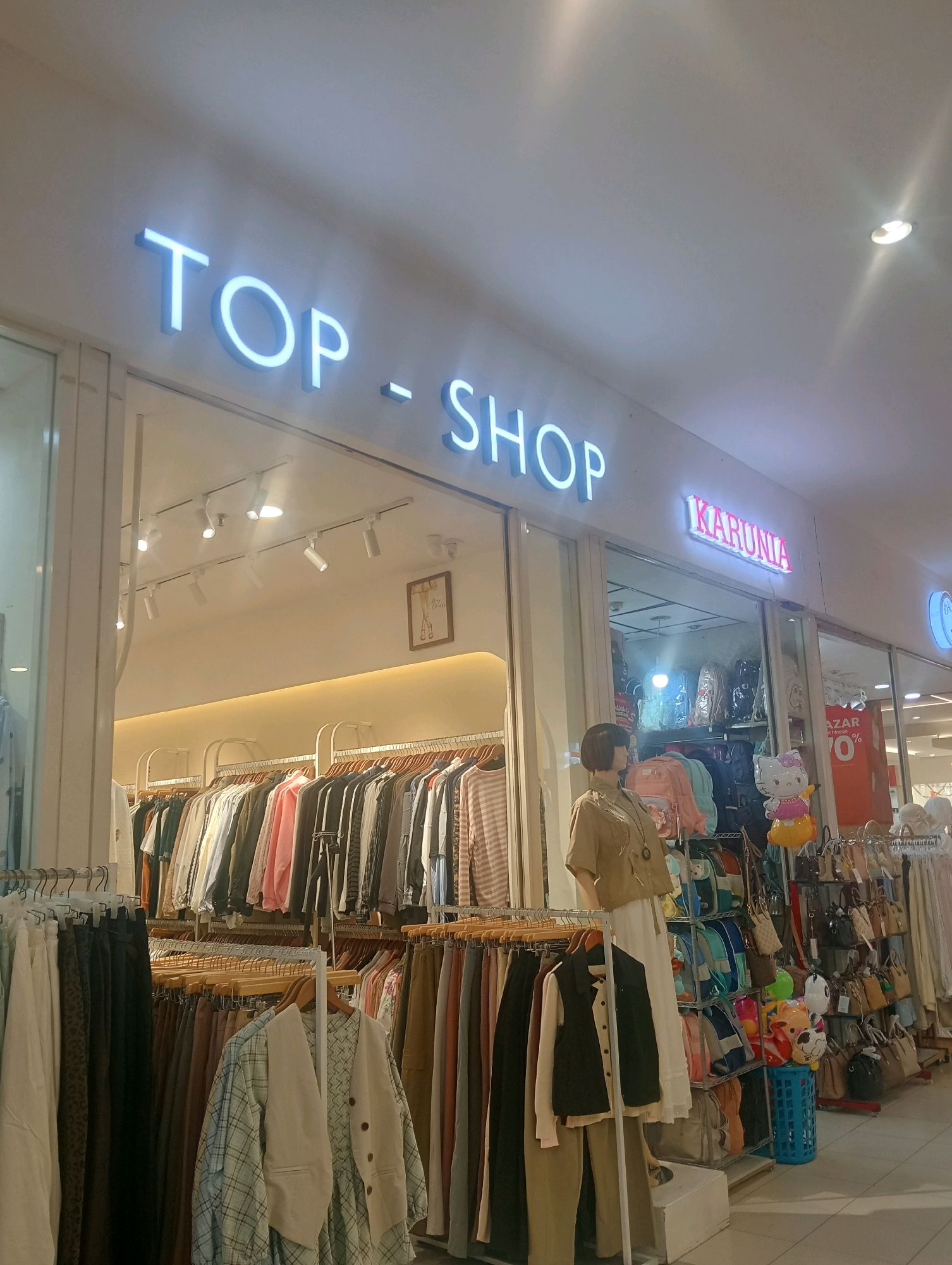 The image depicts the exterior of a clothing store, identified by a prominent neon sign reading "TOP-SHOP". The store appears to be located in a shopping mall or a similar indoor retail environment. To the right of the "TOP-SHOP" sign, another smaller sign reads "KARUNIA" in pink neon. The store's facade is mostly glass, offering a view into the interior where racks of clothing are displayed on hanging rods. A mannequin stands near the entrance, dressed in a neutral-colored outfit. The store is well-lit by overhead track lighting. The overall impression is that of a typical retail space offering women's apparel and accessories. The image context suggests this store is located in Surabaya, Indonesia.