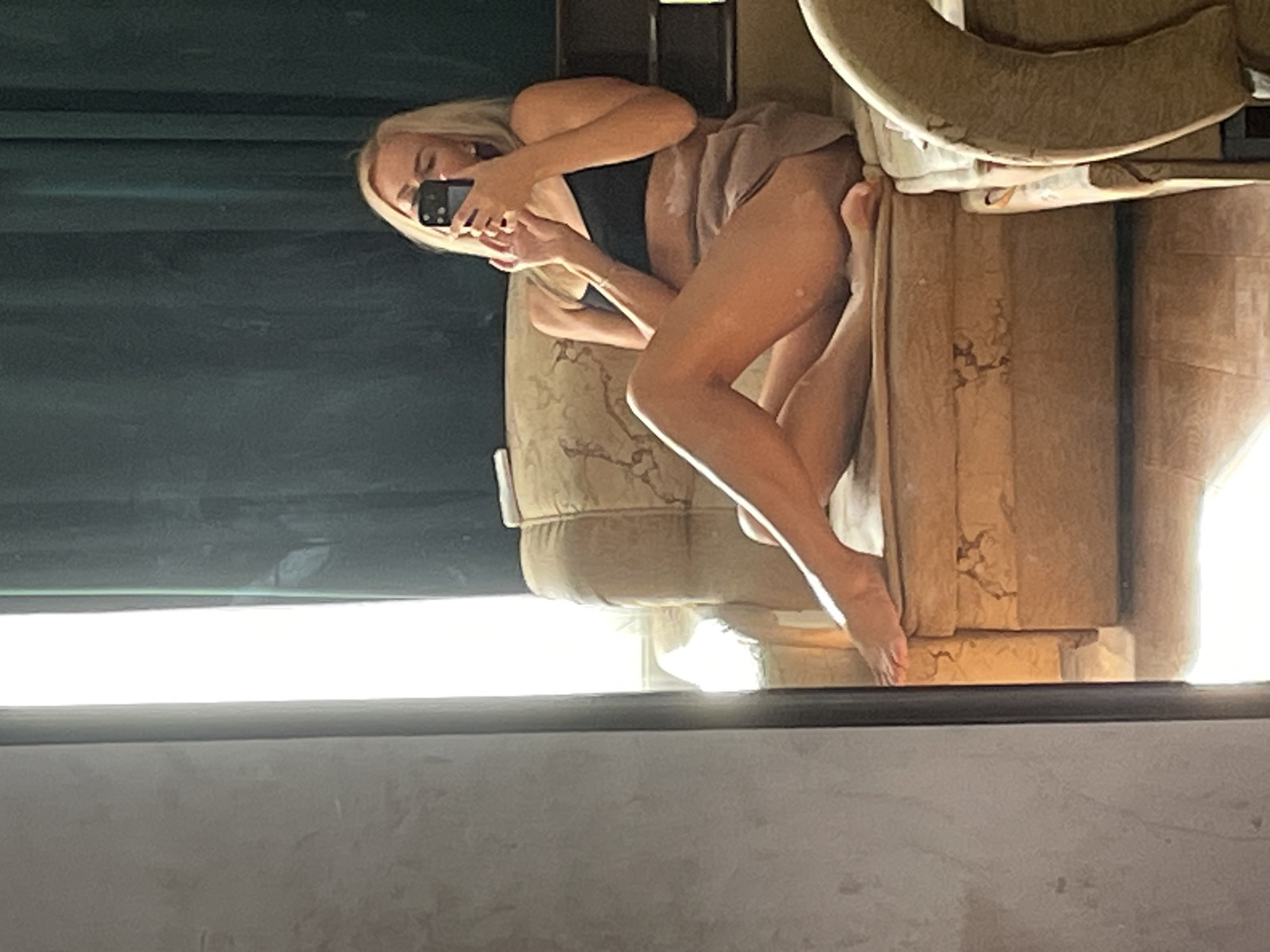 A blonde woman is sitting on a worn, patterned sofa, taking a mirror selfie with a smartphone. She is wearing a black sports bra and light brown shorts. Her legs are crossed, and her feet are bare. The sofa appears to be in a room with a large window covered by dark green curtains, letting in a significant amount of natural light. The room's decor is not clearly visible, but the sofa's condition suggests it might be an older piece of furniture. The image is taken from a slightly low angle, with a wall on the left side of the frame. The setting is domestic, likely a residential apartment. There is no specific indication of the city of Krasnodar, Russia, within the visual information provided in the image.