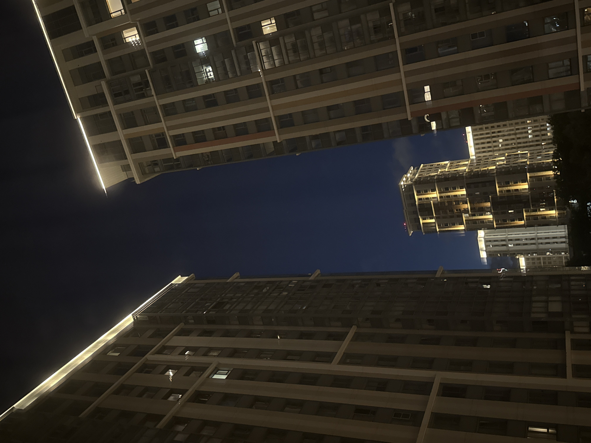 The photograph, taken at night in Khimki, Russia, showcases a modern urban cityscape dominated by tall, illuminated apartment buildings. The perspective is looking upwards between two large, imposing structures, creating a sense of being enclosed within the urban environment.
The building on the left is a dark, multi-story residential complex with numerous windows, some of which are lit, indicating occupancy. A continuous band of warm white light illuminates the top edge of this building, highlighting its modern architectural design and stark contrast against the deep blue night sky.
The building on the right is of similar scale and design, characterized by vertical stripes of beige and darker shades, punctuated by rows of windows. This building also features illuminated windows, suggesting a lively residential presence. Similar to the building on the left, a luminous strip traces its upper perimeter, casting a soft glow.
In the middle ground, a more uniquely designed, illuminated building stands out. It has a stepped, tiered facade, with horizontal bands of light emphasizing its structure. This building appears slightly shorter than the two flanking it, but its distinct lighting pattern makes it a focal point. Behind it, other, less distinct illuminated structures can be seen, contributing to the overall impression of a densely populated urban area.
The sky is a deep, clear indigo, indicative of a cloudless night. The overall impression is one of modern, high-density living, with architecture designed to be visually striking even in the dark. No people are discernible in the image, and the scene is entirely focused on the architectural elements and the ambiance of the city at night. The photograph is a static, objective capture of the urban landscape.