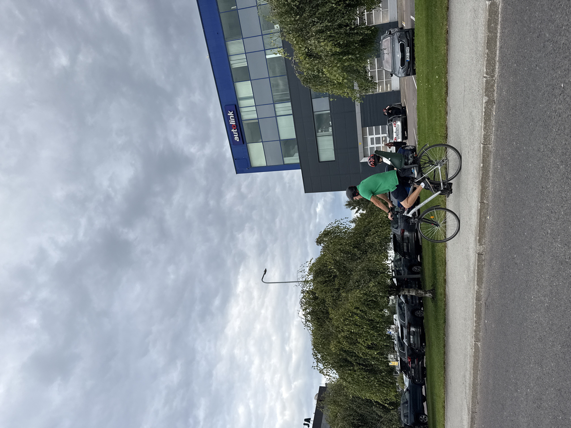 A man in a green t-shirt and dark shorts rides a bicycle with a child in a seat on the back. He wears a black helmet. The man is on a paved path next to a grassy area and a parking lot filled with dark-colored cars. Behind the parking lot is a modern building with a blue upper section and a dark grey lower section. A sign on the blue section reads "autolink." The sky is overcast and grey. The scene appears to be in Tallinn, Estonia, as indicated by the license plates on the cars.