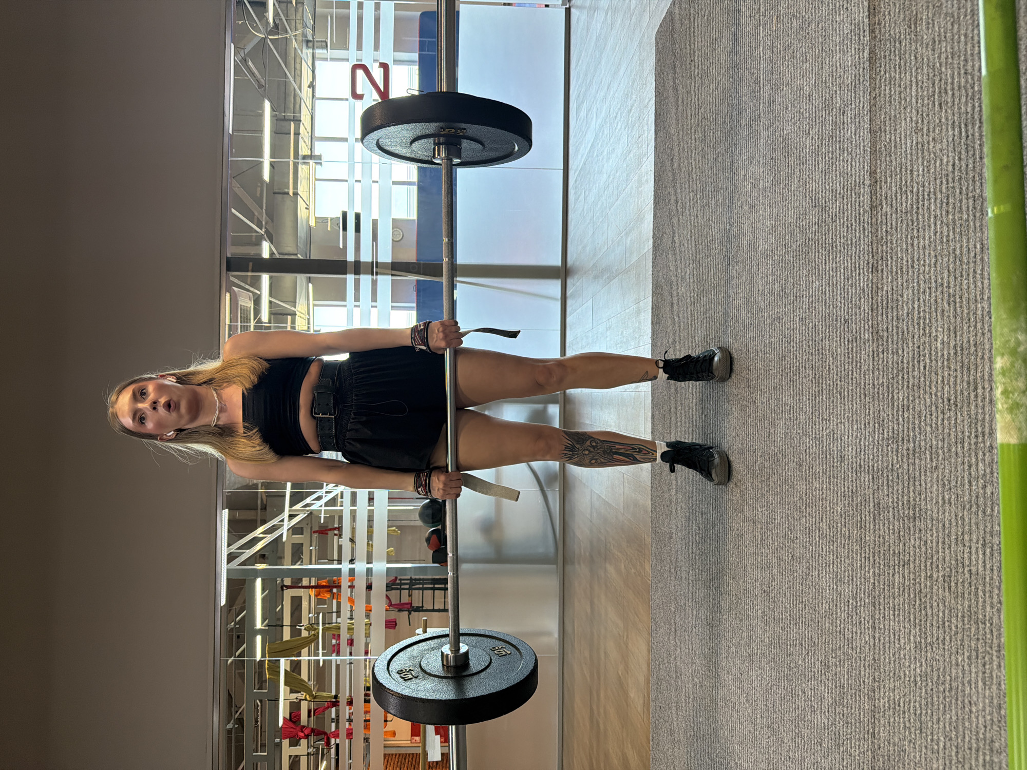 A woman with blonde hair, wearing a black top and black shorts, is lifting a barbell in a gym. She is standing on a grey carpeted floor. Her left leg has a prominent tattoo. The gym has large windows with a view of what appears to be an office building or another part of the gym. The location is likely in Chaikovsky, Russia.