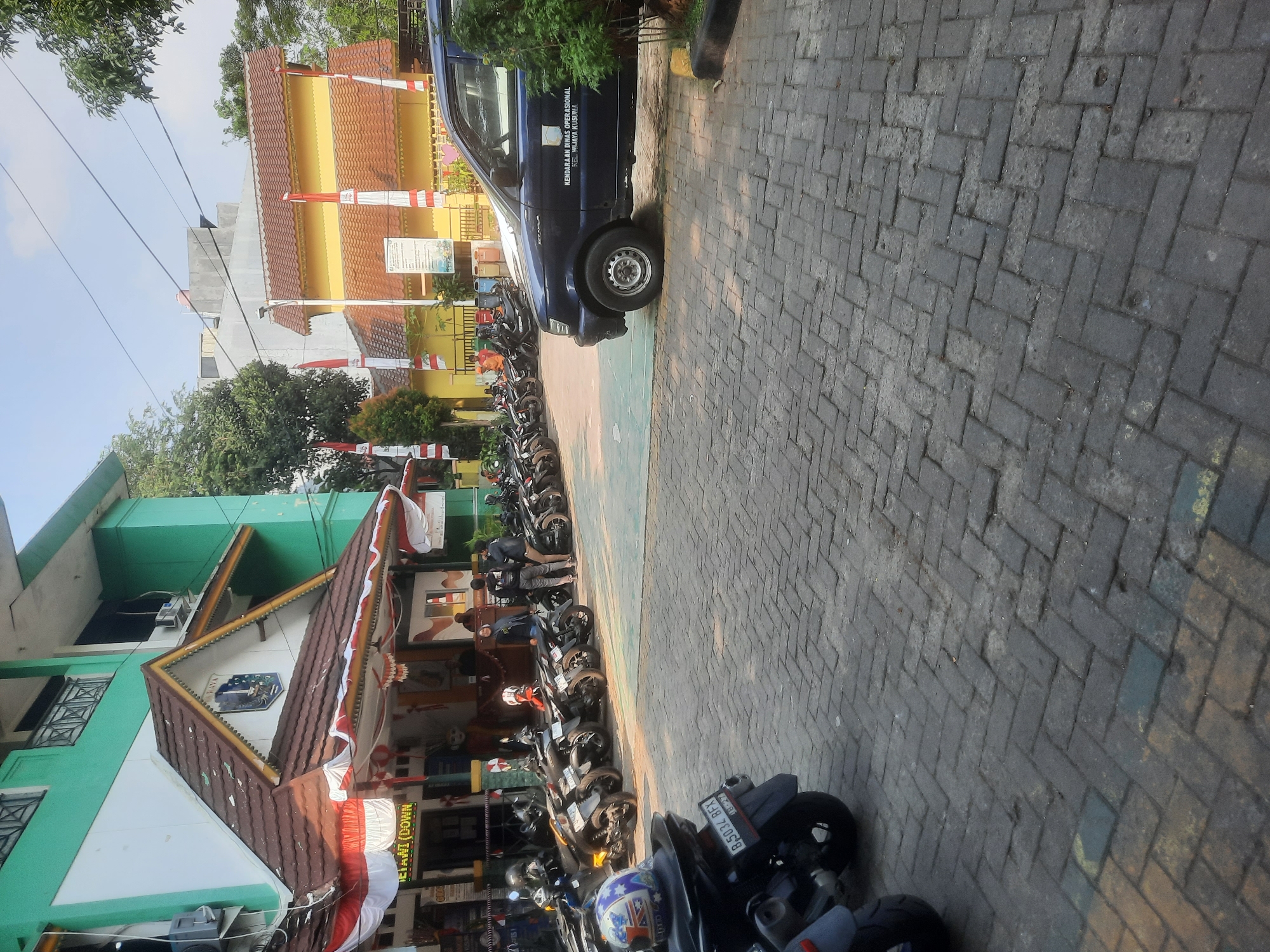 This is an outdoor, eye-level, wide shot of a street in front of a building complex in West Jakarta, Indonesia. The building is a multi-story structure with green and white walls and a brown, tiled roof. The entrance is decorated with red and white Indonesian flags. In front of the building, many motorcycles are parked in rows.
A dark blue SUV with text that reads "KENDARAAN DINAS OPERASIONAL KEL WILAYA KUSUMA" is parked on the right side of the image. There are several people standing and walking around the parked motorcycles. The ground is paved with interlocking bricks. Trees and other buildings are visible in the background. The sky is clear and blue.