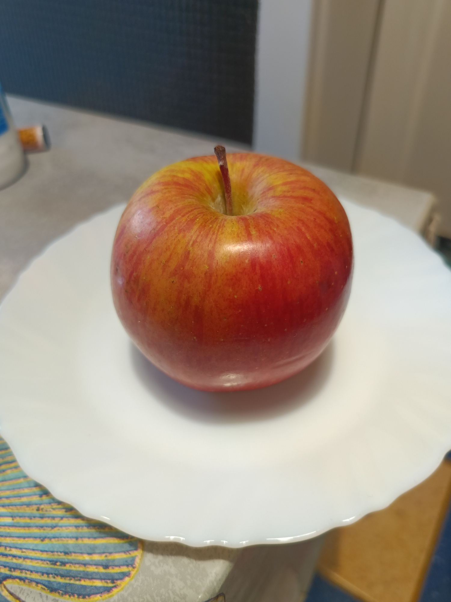 A single, ripe apple with red and yellow streaks sits on a white, scalloped plate. The plate is positioned on a table, which appears to be a kitchen counter, as suggested by the blurred background that includes a tiled surface and what might be a cupboard or wall. The lighting is natural, likely from a window, casting a soft shadow from the apple onto the plate. The scene is static and appears to be an indoor, domestic setting. No specific location within Varna, Bulgaria can be determined from the image.