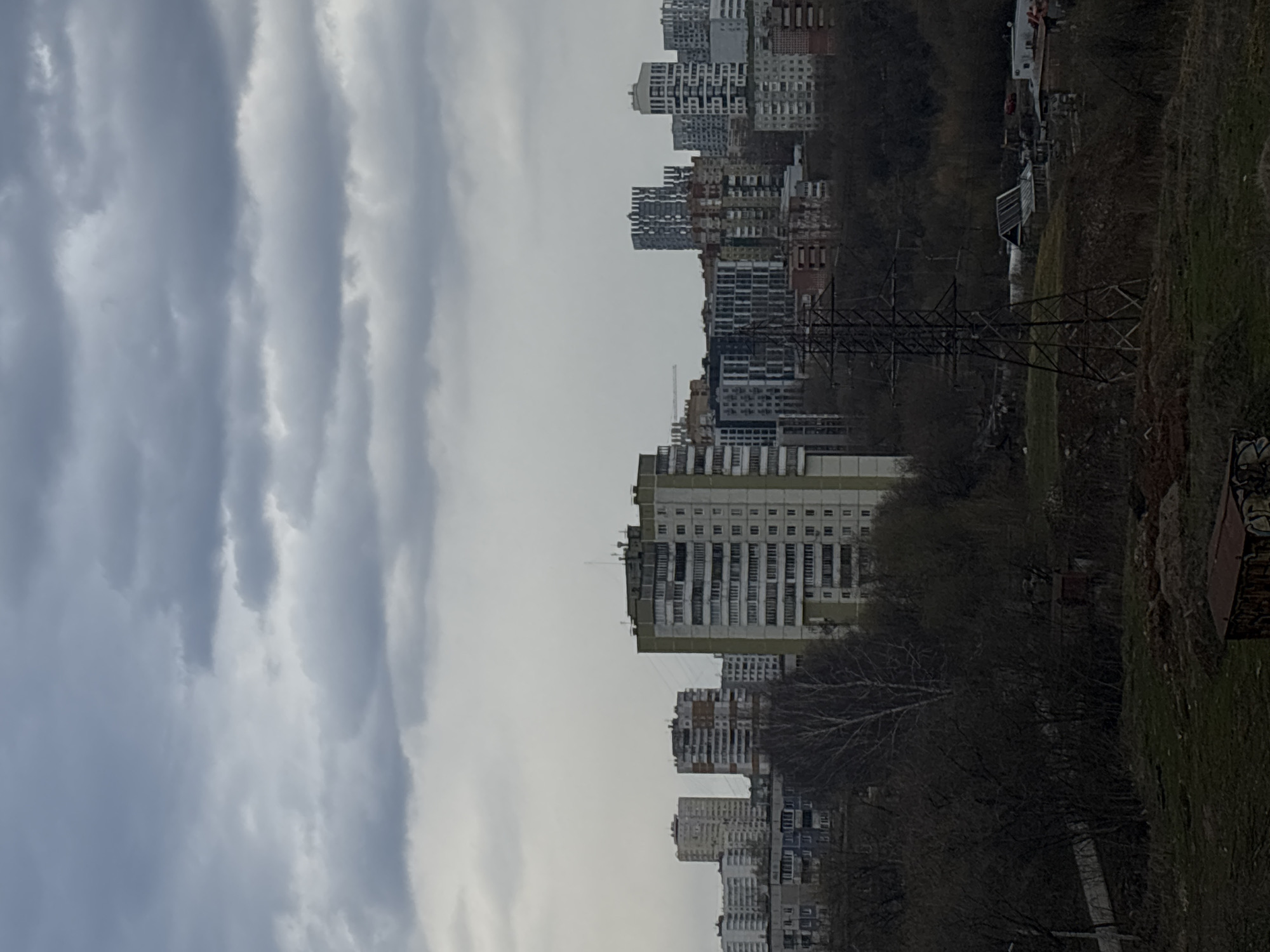 This image captures an urban landscape under a heavily overcast sky, suggestive of a cloudy day. The scene is dominated by a dense array of multi-story residential buildings, likely apartment blocks, stretching across the middle ground. These structures vary in design, with some appearing older and more uniform, featuring numerous windows and balconies, while others might be newer or under construction, indicated by the fainter appearance of a construction crane in the distance.
The foreground and right side of the image (when correctly oriented) feature a sloped, natural area with bare trees, hinting at late autumn or early spring. A prominent, dark metal lattice power transmission tower stands in the mid-ground, adding an industrial element to the urban sprawl. The ground is covered with patchy brown and green grass. On the far right, a small, box-like structure with graffiti is visible. The sky occupies a significant portion of the frame, filled with textured, light grey clouds that create a diffused, ambient light over the entire scene. There are no people visible, and no specific activities are taking place. The overall impression is that of a typical urban periphery in Perm, Russia, during a non-sunny day. No discernible text is visible in the image.
