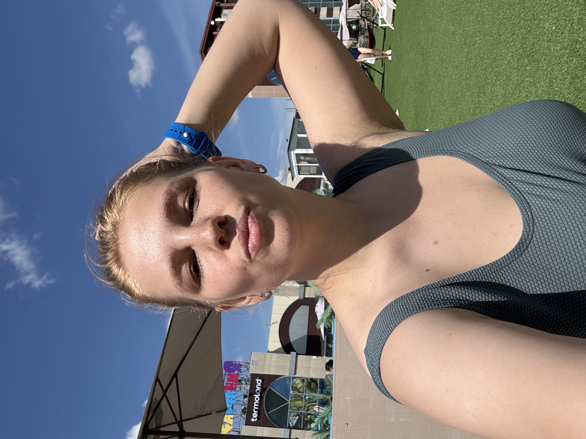 In an outdoor, rooftop setting, a young woman with blonde hair pulled back poses for a selfie against a bright blue sky with scattered clouds. She is wearing a teal-colored, textured swimsuit and a blue wristband. Her right arm is raised, with her hand resting on her head. In the background, a building with signage that reads "termoland" is visible, along with a large umbrella, lounge chairs, and green turf. The scene suggests a sunny day, possibly at a water park or resort.
