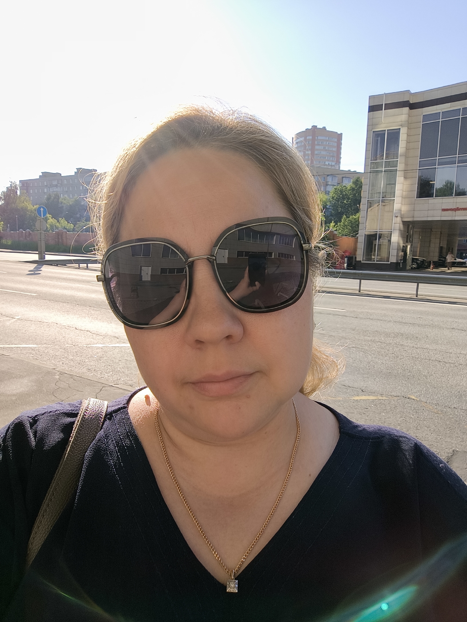 A woman wearing large sunglasses and a dark V-neck top with a gold chain necklace is taking a selfie outdoors in Moscow, Russia. She is standing on a sidewalk next to a street with a building in the background. The building appears to be a modern office or retail building with large windows. Sunlight is visible in the image, casting shadows and creating lens flare. Traffic is present on the street in the background.