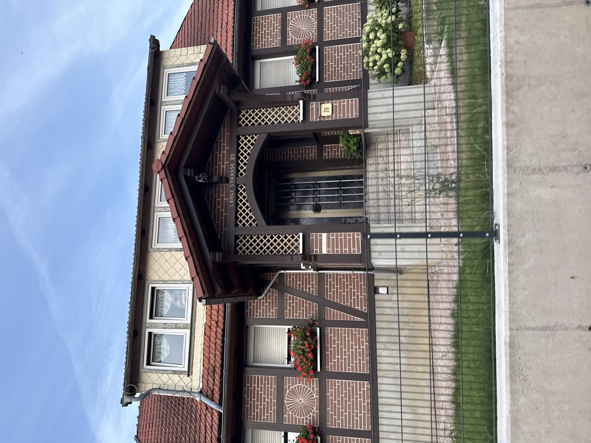 The image shows a two-story house with a red tile roof, typical of German architecture. The facade is constructed with dark brown timber framing against a reddish-brown brick background, a style known as Fachwerkhaus. The windows on the upper floor are small and rectangular, while the ground floor features larger windows with white shutters and flower boxes filled with red flowers. A prominent entryway features a dark brown wooden porch with a lattice design. Above the entrance, a sign reads "LANGE STRASSE 39," indicating the street and house number. A small yellow house number sign, also reading "39," is affixed to the wall to the right of the entrance. The house is surrounded by a metal fence and a concrete retaining wall. In front of the house, a small patch of green grass is visible, and to the right of the entryway, a large bush with white flowers is planted in a pot. The sky is blue with some wispy clouds, suggesting it is daytime. The image appears to be taken from a slightly elevated position, possibly from across a street or a lower part of the property. The scene depicts a residential building in a suburban or semi-rural setting, consistent with a town like Bad Sachsa, Germany, known for its charming half-timbered houses.