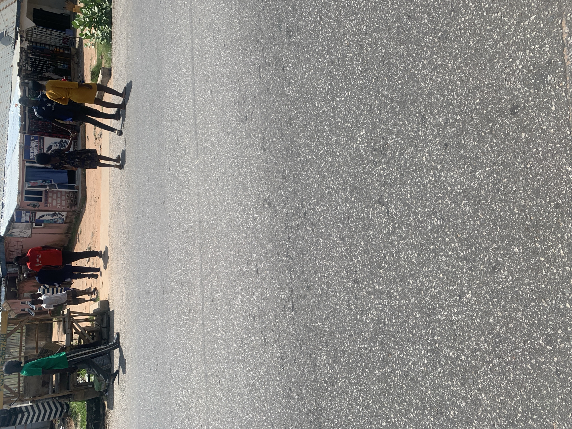 The image depicts a street scene in Jalingo, Nigeria. Several people are visible standing or walking along the side of a paved road.
In the foreground, on the left side of the image, a person wearing a green shirt and black pants is standing next to a wooden structure. Further up the road, a group of four people are gathered. One person is wearing a red shirt, another a dark blue shirt with white stripes, and a third a yellow shirt. The fourth person is wearing a dark shirt and dark shorts. They appear to be casually interacting. Behind them, there are some buildings with storefronts, suggesting a commercial area. One sign visible reads "LEGIT DIAGNOSTIC LABORATORY".
The scene is outdoors, under what appears to be daylight, likely mid-morning or afternoon given the shadows. The weather seems clear and sunny. The overall atmosphere appears casual and everyday. The road is asphalt, with some texture visible. The image is oriented vertically, with the subjects appearing in the left portion of the frame.