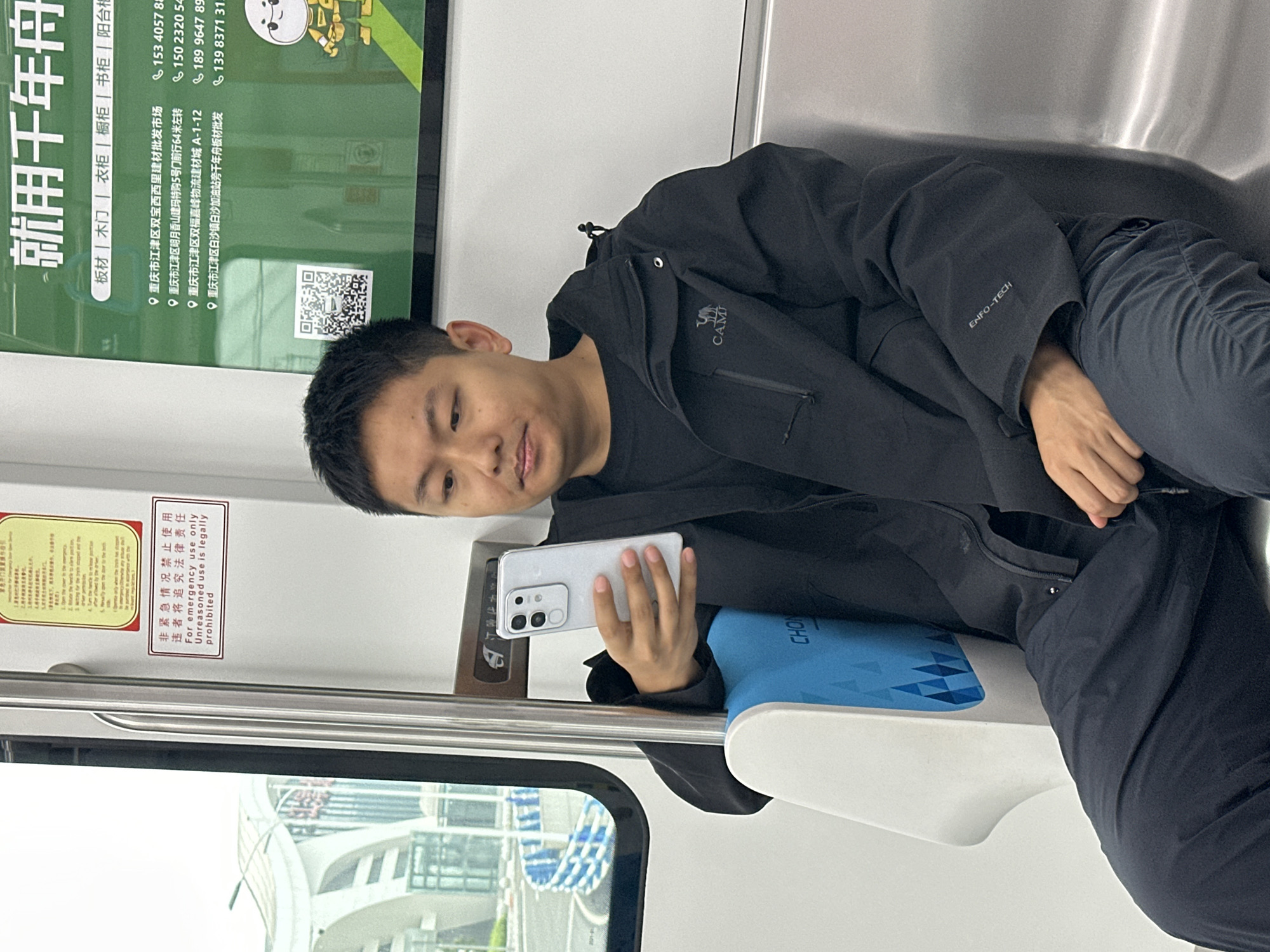 A young man with short dark hair, dressed in a black jacket and dark pants, is seated inside what appears to be a modern train or subway car. He is looking down at a light-colored smartphone he holds in his right hand, seemingly engrossed in its content. The man has a calm and focused expression.
The interior of the train is clean and bright, with large windows providing a glimpse of the outside world, showing structures that resemble urban architecture. A blue and white patterned cushion is visible beneath him.
Several advertisements and signs are displayed on the wall of the train car. A large green advertisement for a brand named "千年舟" (Qiannianzhou) lists products like "板材 | 木门 | 衣柜 | 橱柜 | 书柜 | 阳台柜" (Board | Wooden door | Wardrobe | Cabinet | Bookshelf | Balcony cabinet). Below this, multiple addresses in Chongqing's Jiangjin District are listed, including locations in "双宝西西里建材批发市场" (Shuangbao Xixili Building Materials Wholesale Market), "明月香山建玛特购" (Mingyue Xiangshan Jianmate Shopping), "双福嘉峰物流建材城" (Shuangfu Jiafeng Logistics Building Materials City), and near "白沙加油站" (Baisha Gas Station), along with various phone numbers. This confirms the setting is in or around Jiangjin, China.
Another sign, white with red text, warns in both Chinese and English: "非紧急情况禁止使用 违者将追究法律责任 For emergency use only Unreasoned use is legally prohibited." This likely refers to an emergency brake or exit mechanism. The man's black jacket features a small camel logo with the text "CAMI" and the word "ENFO-TECH" embroidered on the sleeve. The overall lighting suggests it is daytime.