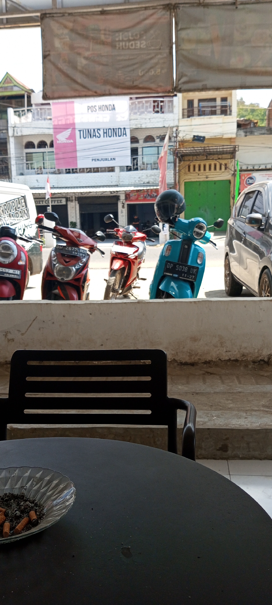 The image depicts an outdoor scene likely in Lempa, Indonesia. In the foreground, a black circular table is visible with an ashtray filled with cigarette butts. Behind the table, a black metal chair with horizontal slats is positioned. In the middle ground, several motorbikes are parked, including a red scooter on the left, a red motorbike in the center, and a teal motorbike with a black helmet on its handlebar to the right. The teal motorbike has a license plate reading "DP 5038 UK". A grey car is parked behind the teal motorbike. In the background, a building with multiple levels and windows is visible. A large banner with "POS HONDA TUNAS HONDA" and a Honda logo is hanging from the building. Other banners and signs are also present, including one with partially visible text. The environment suggests a commercial area, possibly a dealership or a public space with shops. The lighting indicates it is daytime.
