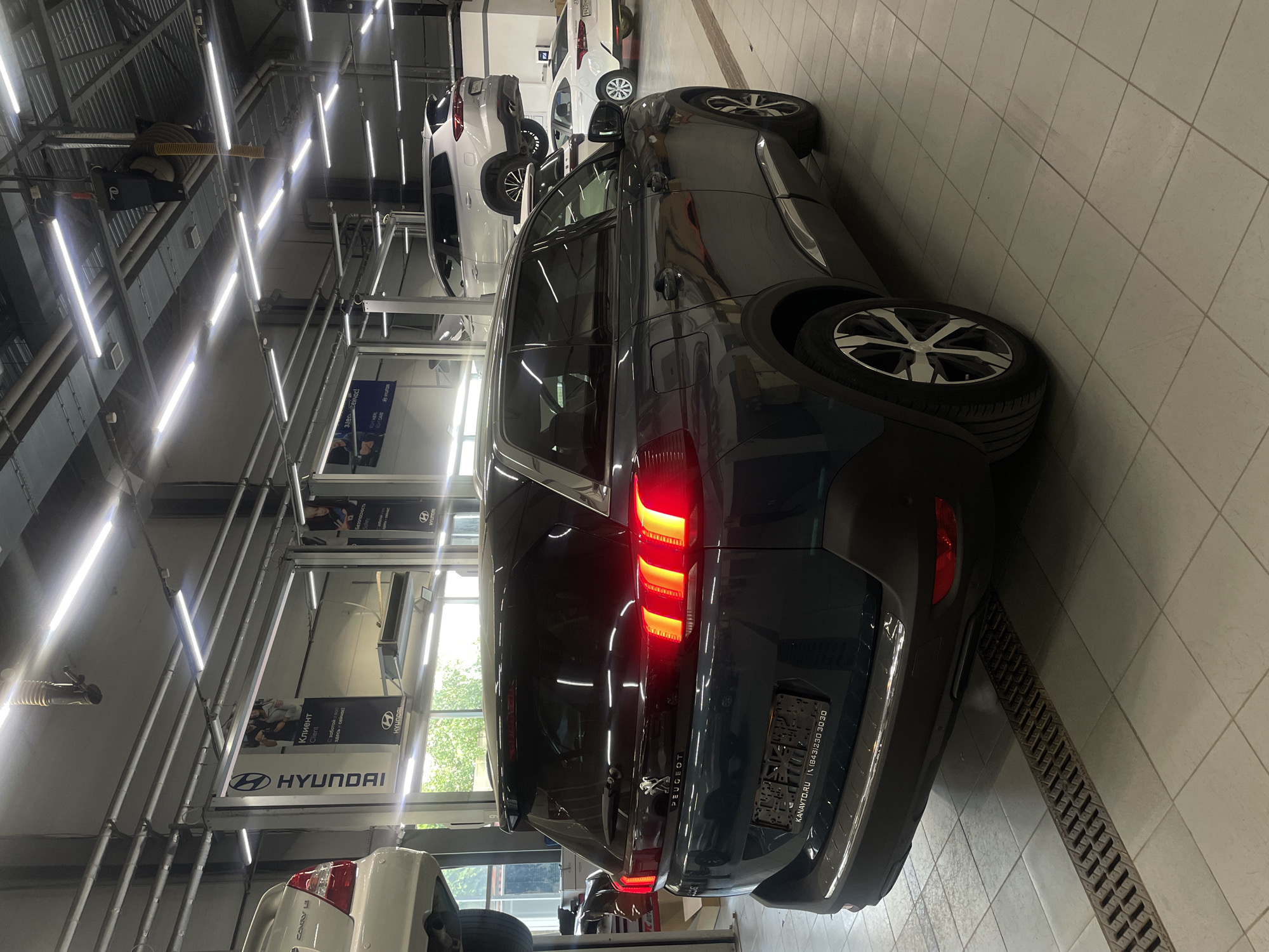 The image depicts the interior of a modern car service center in Kazan, Russia. The primary subject is a dark-colored Peugeot SUV parked on the floor, facing away from the camera. Its brake lights are illuminated. In the background, several other vehicles are visible: a white sedan is parked on the right, and a silver SUV is elevated on a car lift to the left. Above the vehicles, the service center's ceiling is equipped with bright fluorescent lighting and various mechanical components. Banners advertising Hyundai are visible on the walls. The floor is tiled and features a drainage grate in front of the Peugeot. The overall environment suggests a functional and well-lit automotive repair or dealership facility.