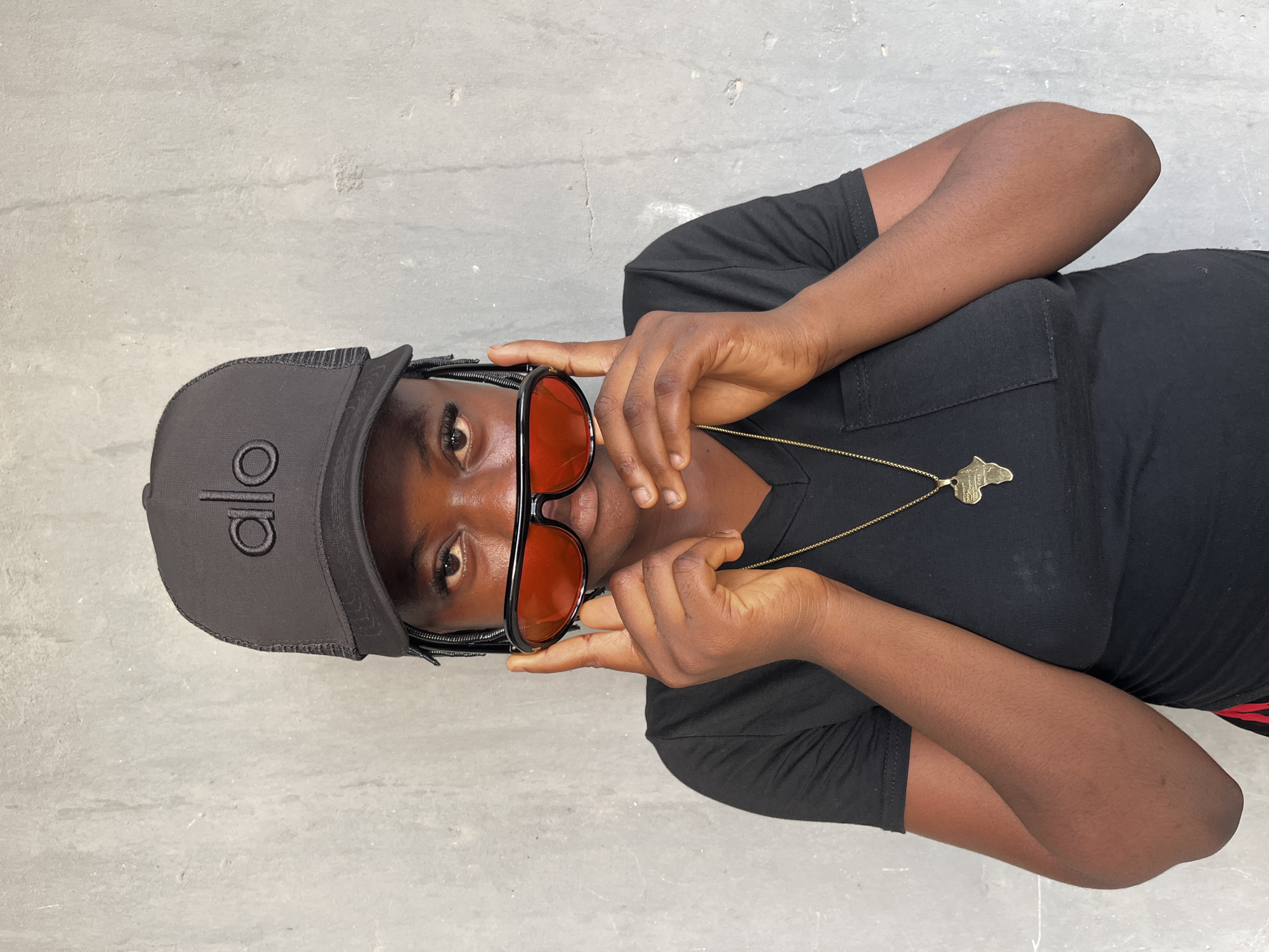 A woman is centered in the frame, wearing a black t-shirt and a black baseball cap with the logo "alo" on the front. She is also wearing sunglasses with orange-tinted lenses and a gold necklace with an African continent pendant. She is holding the sunglasses with her hands, partially obscuring her face. The background is a light grey, textured surface, likely a concrete floor or wall. The image was taken in Badagry, Nigeria.