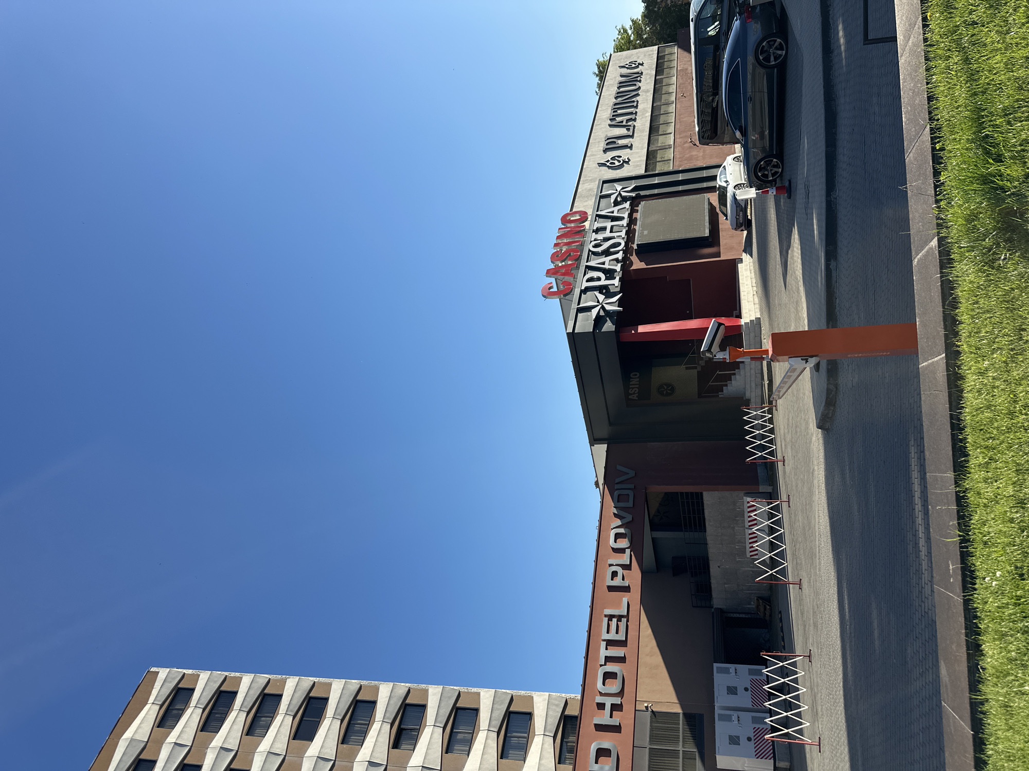 The image shows the facade of the "RD Hotel Plovdiv" and "Casino Pasha & Platinum" in Plovdiv, Bulgaria. The hotel has a multi-story building with a patterned exterior, while the casino is attached to the hotel and displays red signage. Two cars are parked along the pavement, and a stretch of green grass lines the right edge of the image. A blue sky fills the upper left portion.