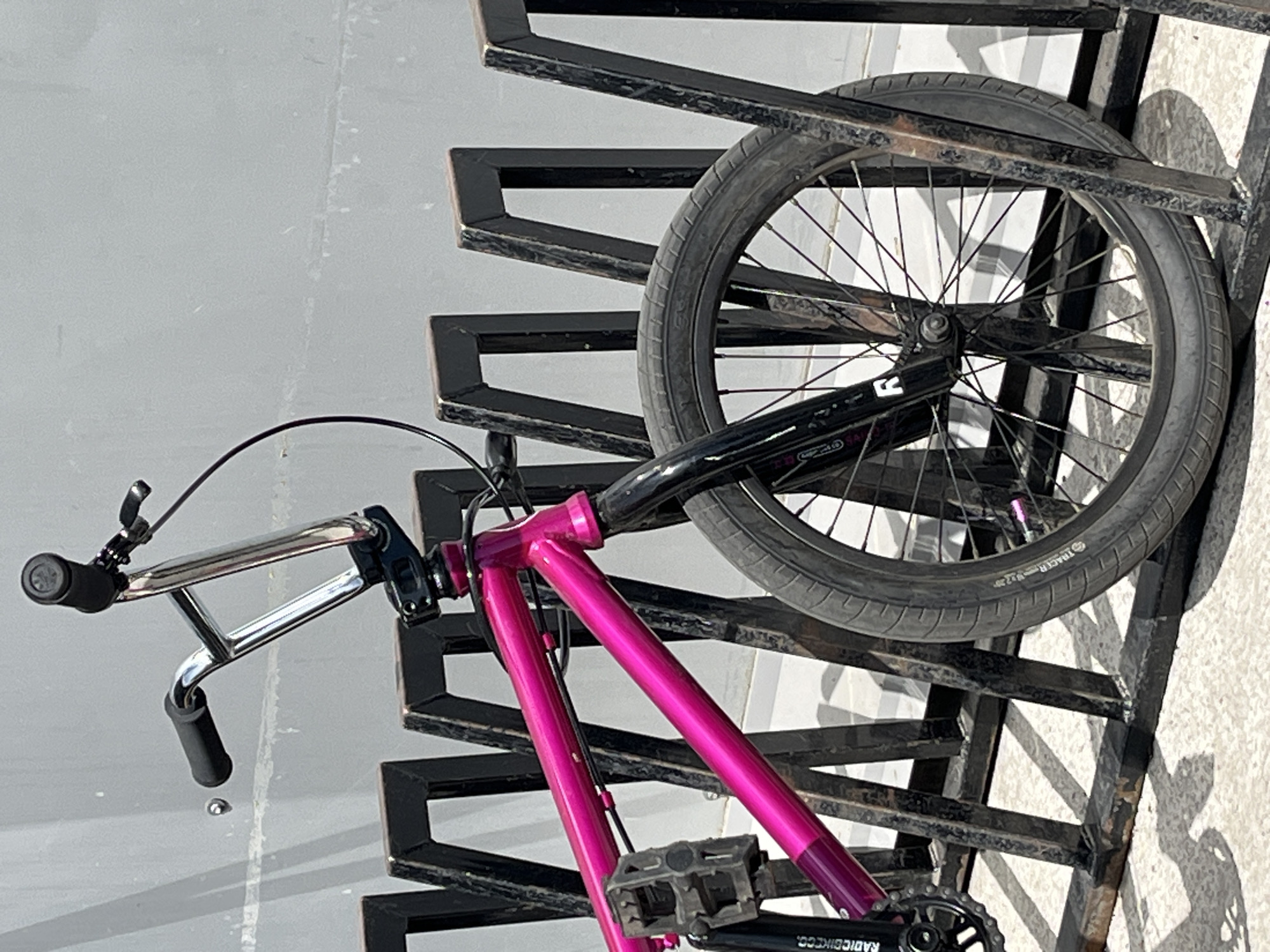 A hot pink and black BMX bike is locked to a black metal bike rack on a grey surface. The bike's front wheel is secured within the rack. The frame of the bike is hot pink, while the handlebars, forks, and various other parts are black. The tire is a black "Tracer 2.25" brand. The pedals and chainring are visible. Based on the location information provided, this image was taken in Krasnoyarsk, Russia, although there are no specific location cues visible in the image.