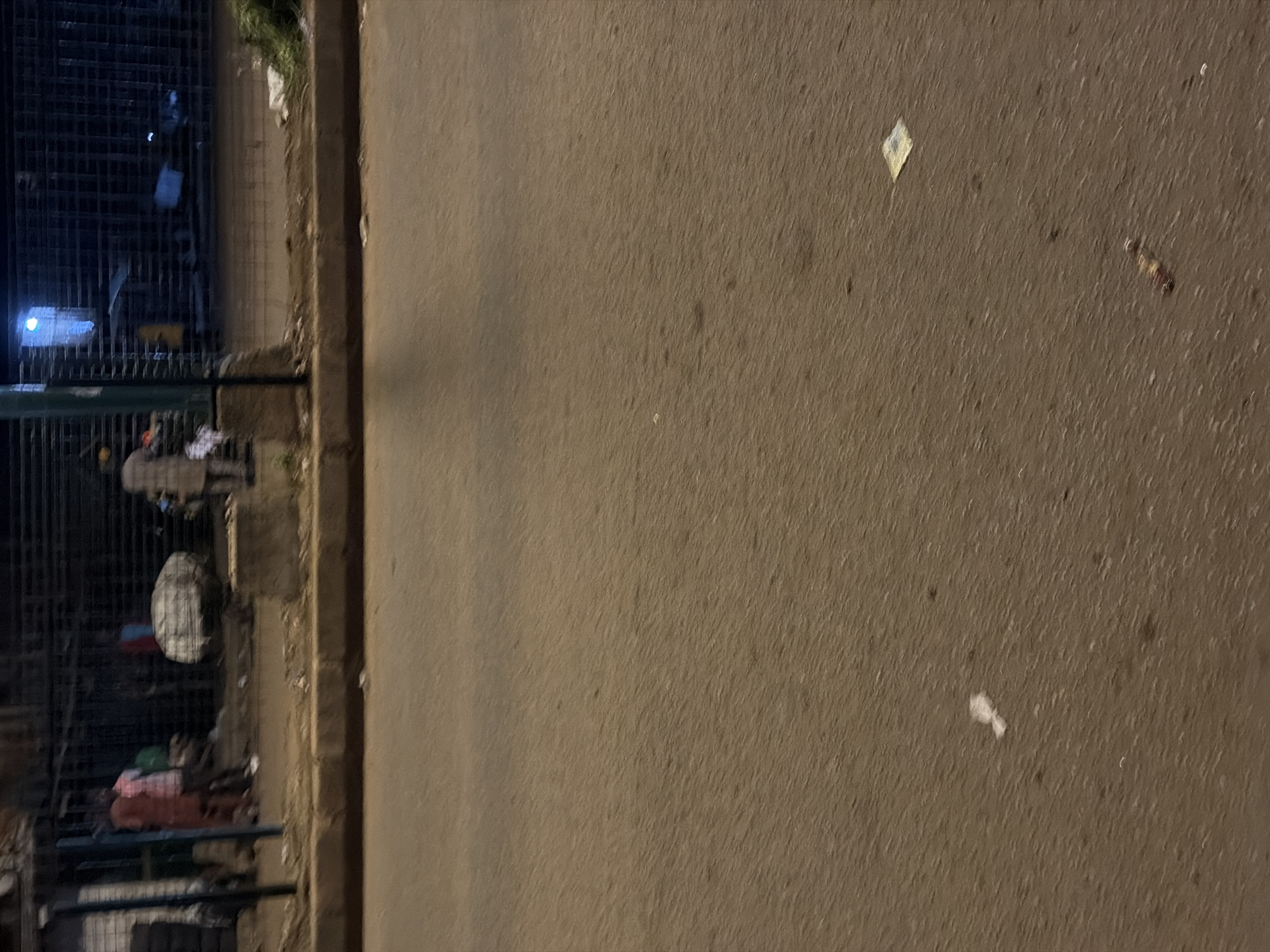 This is a street-level, medium-distance, eye-level, still photograph of a street in Bukuru, Nigeria. The scene is composed of two distinct planes: the lower half of the image is dominated by a paved road surface, while the upper half depicts the edge of a sidewalk with an overhang structure and pedestrians. The road is a dull, reddish-brown asphalt, with visible texture and a scattering of debris, including a small white wad of material on the left, a yellowed square object, possibly currency or a receipt, and a thin, reddish object, possibly a piece of food or debris, on the right.
The upper section of the image reveals a concrete sidewalk with a raised curb separating it from the road. Behind the curb is an area with a metal grate structure, suggestive of a shopfront or covered market. Several individuals are present in this area. On the left, two people are standing close together; one appears to be wearing a reddish-brown outfit, and the other a pink and white patterned shirt. Further back, a figure in a light-colored garment with darker patterns is standing near a large, irregularly shaped bundle covered in a white material. To the right, more of the grate structure is visible, with another person standing near it. A bright blue light emanates from within the structure on the far right. Green foliage is visible peeking out from the right side of the frame, near the grate. The lighting suggests it is either dusk or nighttime, with artificial lights from within the structures casting shadows. The overall impression is a typical urban street scene in Nigeria, with signs of commercial activity and pedestrian presence.