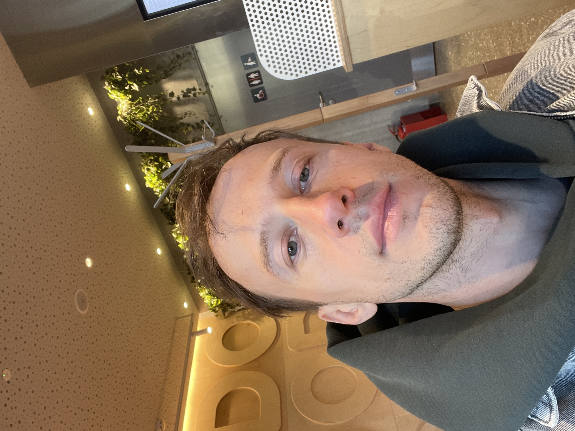 An indoor, vertically oriented selfie captures a man from the chest up, looking directly at the camera with a neutral expression. He has short, light brown hair, blue-green eyes, and light stubble on his chin and jawline. He is wearing a dark green hooded top or jacket over what appears to be blue denim clothing.
The background suggests a modern public space, possibly a cafe or restaurant, in Murino, Russia. To the man's left, a light-colored, textured wall with small perforations is visible, illuminated by several recessed circular lights. Further left and behind him, a wall is adorned with artificial or live green vines, suggesting natural decor.
Above the man's head, part of a metallic structure with a white perforated panel is visible, next to three small pictogram signs. These signs depict a standing figure, a sitting figure, and a disabled access icon, commonly found on restroom doors. Below these, a small red box, possibly a fire alarm or electrical component, is mounted. Wooden panels and structures are also part of the background, potentially forming a counter or table. To the man's right, large, light-colored embossed letters reading "DO" are partially visible on a wall, suggesting part of a brand name or decorative signage. The lighting in the scene is artificial and warm.