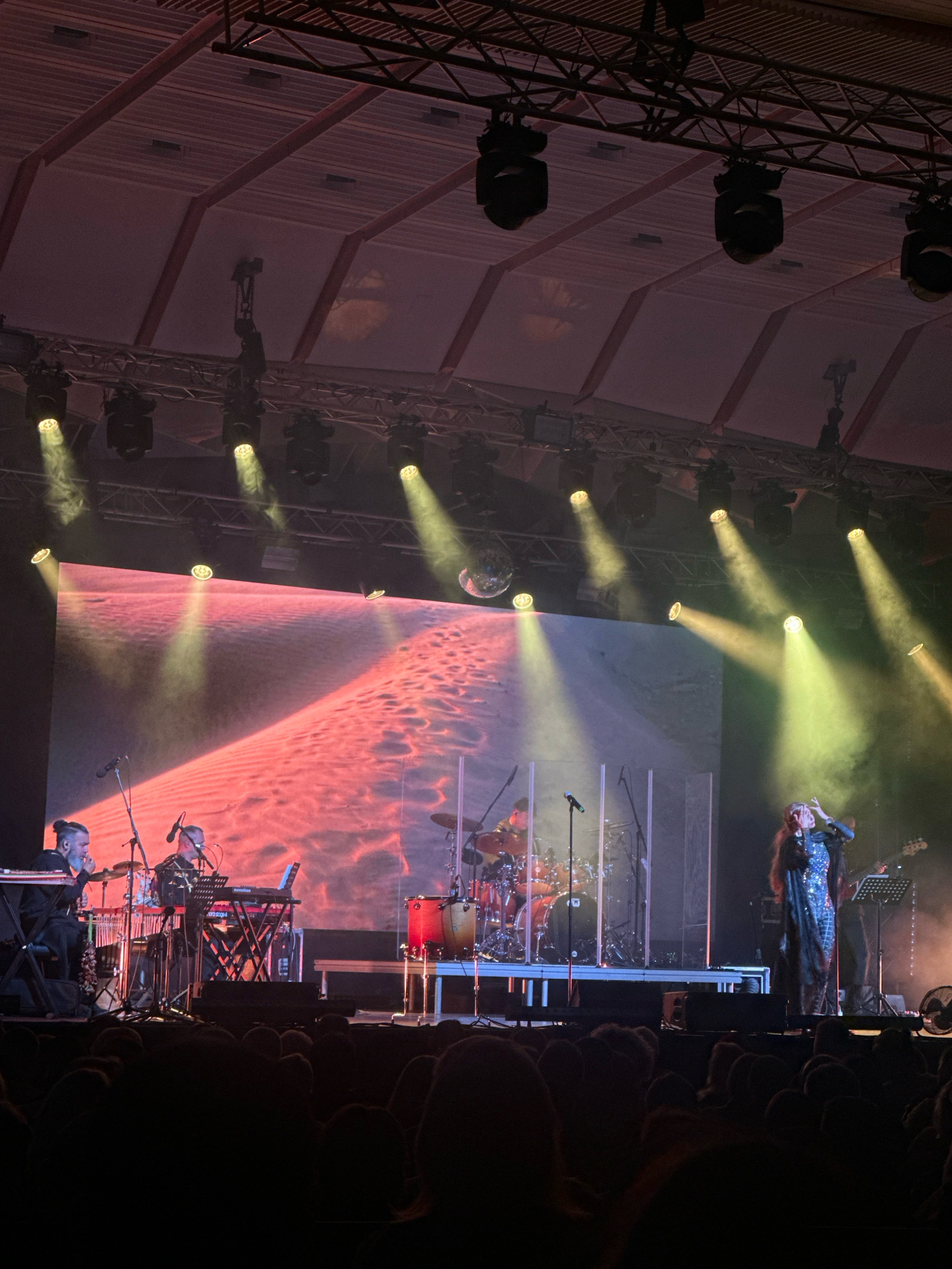 A live concert is taking place in an indoor venue. A band is performing on a stage with a large screen displaying a red and pink abstract background with a sandy texture. In the foreground, to the left, two musicians are playing keyboards. One musician, with dark hair pulled back in a bun, is seated and looking down at his instrument. The other musician is also seated and appears to be singing into a microphone. Behind them, a drummer is seated at a drum kit, with red and silver drums. To the right of the stage, a female singer in a sparkly black and silver outfit is singing into a microphone, gesturing with her hands. Another musician is visible in the background, playing a guitar. The stage is lit by several spotlights, casting beams of light and creating a smoky atmosphere. The audience is seated in the dark in the foreground, with their backs to the camera. The venue appears to be a concert hall or arena with a suspended ceiling and lighting rig. This scene is likely taking place at the Dzintari Concert Hall in Jūrmala, Latvia, given the context of live musical performances in that city.