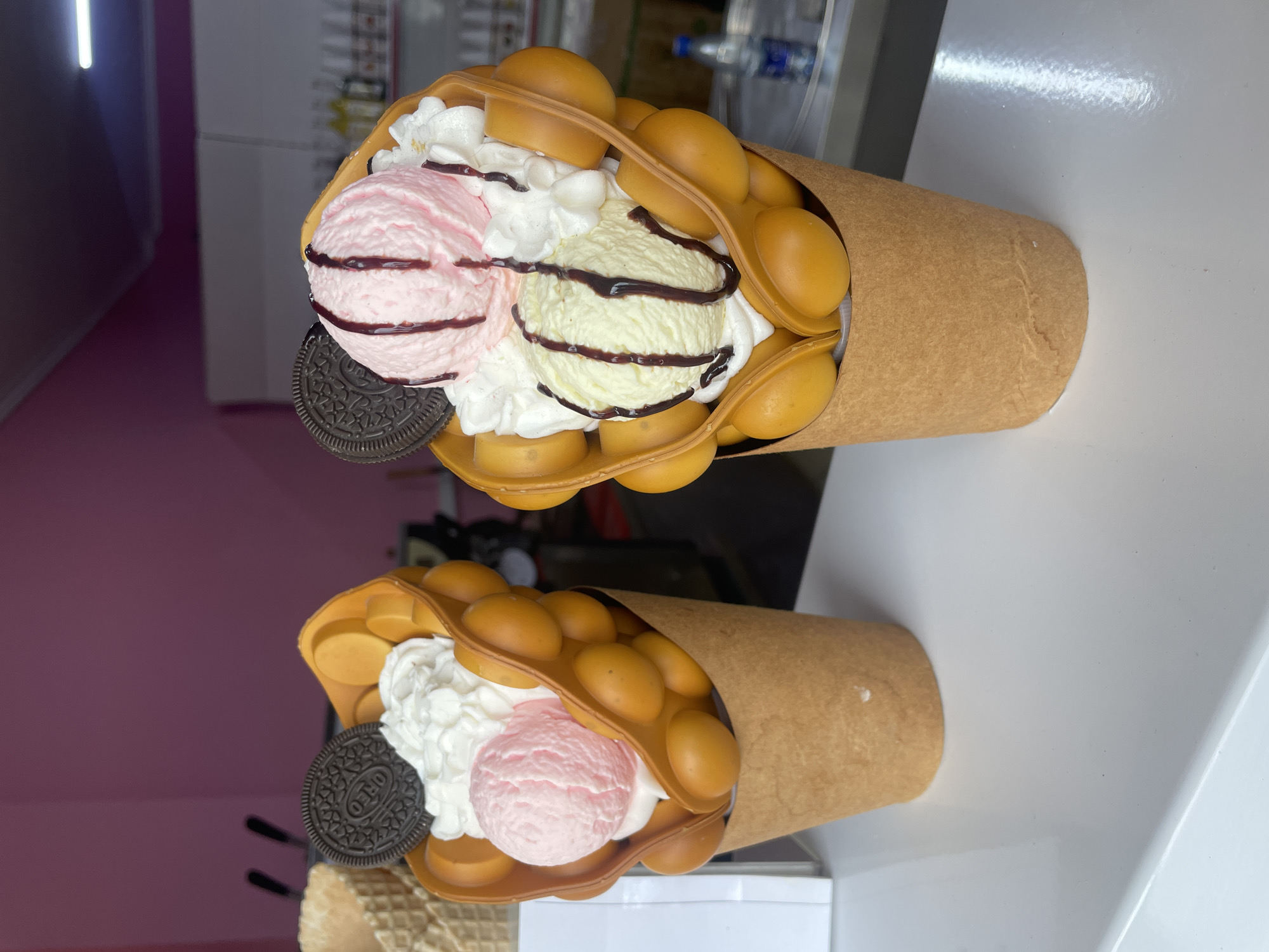 The image shows two bubble waffle cones filled with ice cream, whipped cream, chocolate syrup, and an Oreo cookie. The waffle cones are placed on a white countertop. The background is blurry, but shows a water bottle. The location is unknown but does not appear to be in Olginka, Russia.
