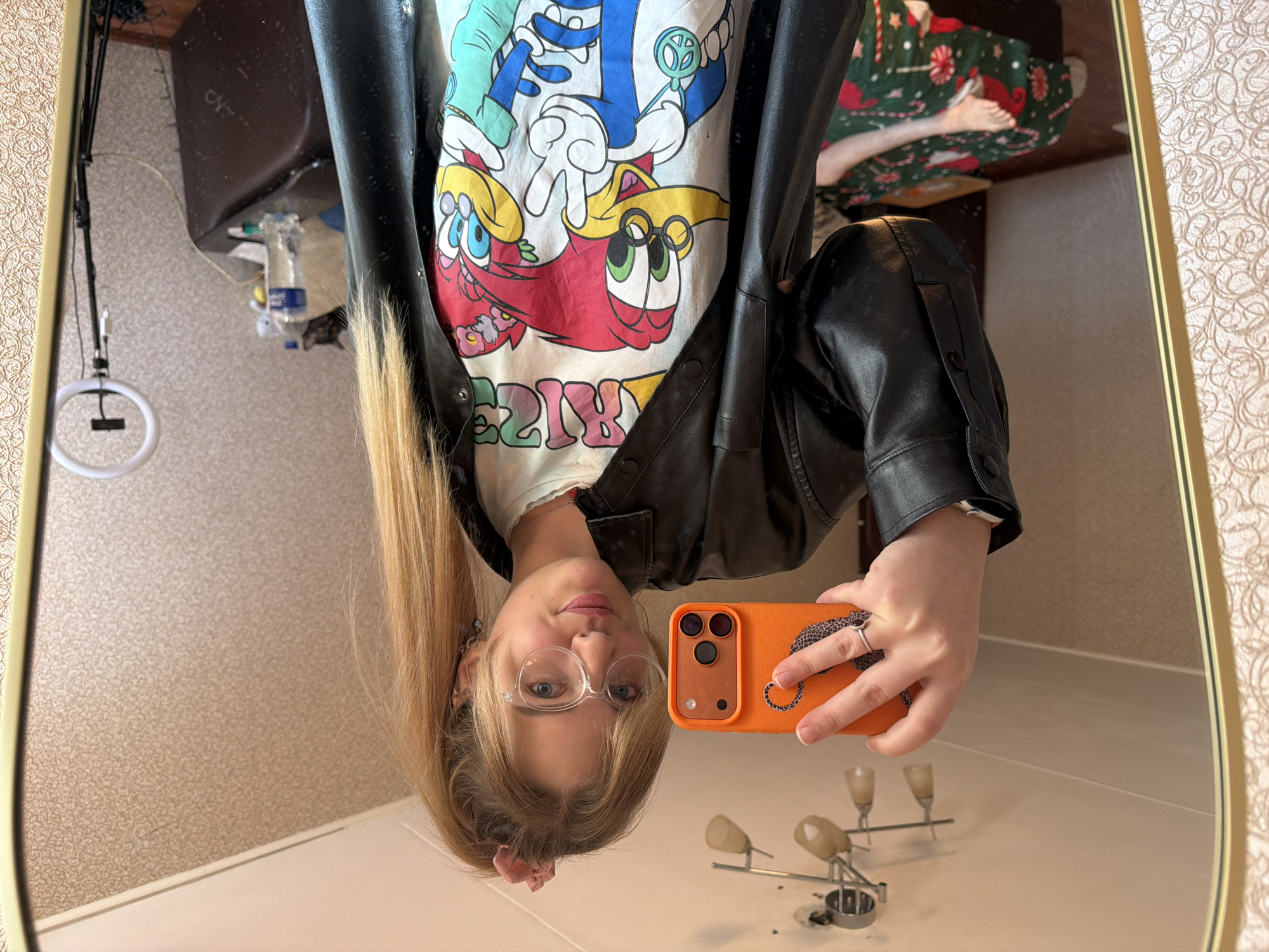 A young woman with blonde hair tied back is taking a selfie in a mirror. She is wearing glasses, a graphic tee shirt featuring cartoon characters, and a black leather jacket. She is holding an orange smartphone in her right hand. The mirror reflects a portion of a bathroom or similar space with patterned wallpaper and a white countertop. A sink fixture and some toiletries are visible in the reflection. A lamp with a circular light is hanging on the side. A bed with Christmas sheets is in the background. It is not possible to determine the location as Naberezhnye Chelny, Russia, from the image alone.