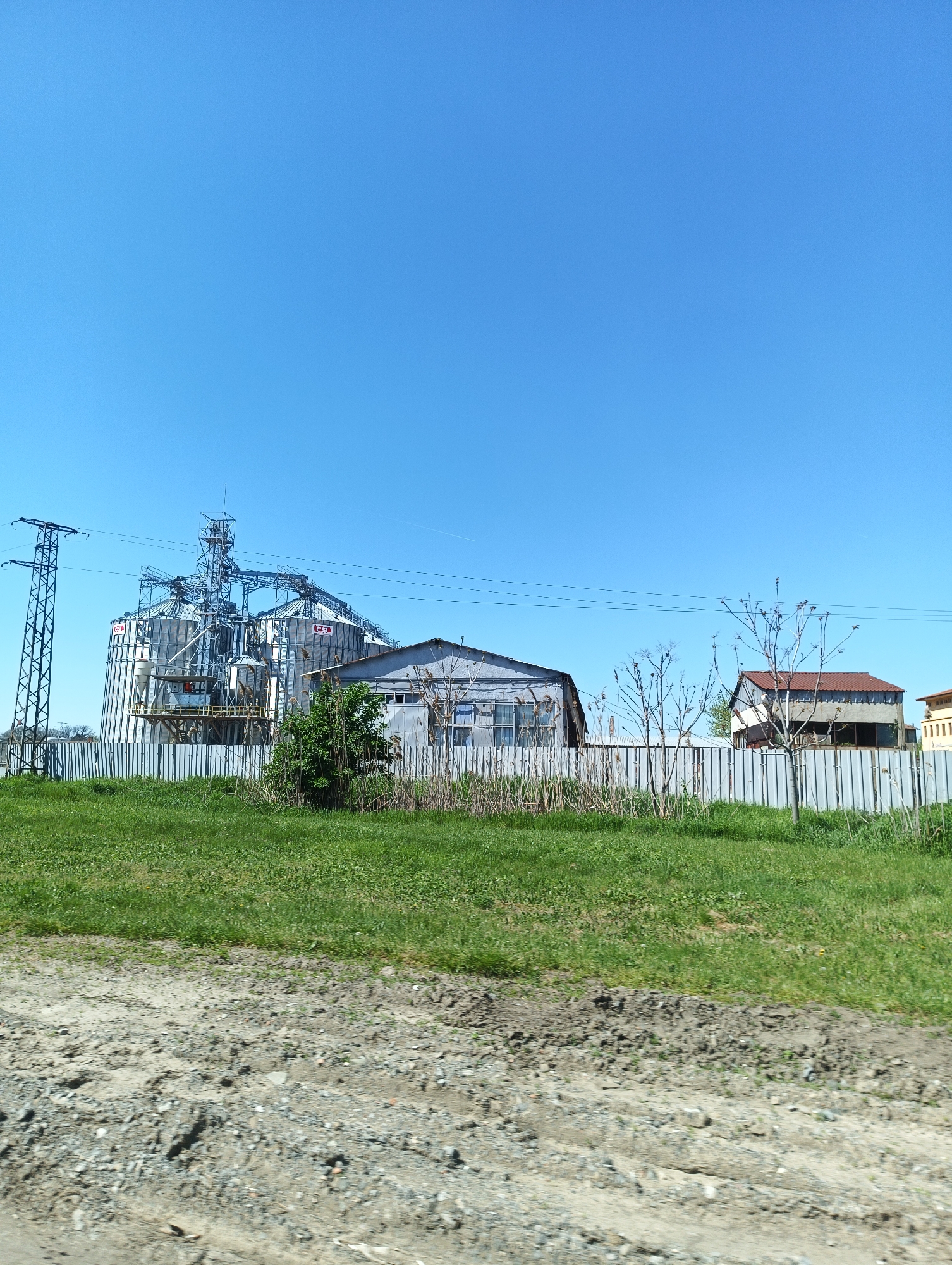 The image shows a rural scene in Straldzha, Bulgaria. In the background, there are three large silver silos, next to a warehouse, an electrical transmission tower, utility lines, and a one-story home with a red tile roof. A faded white corrugated metal fence runs along the property line, with grasses and small shrubs. In the foreground there's a strip of green grassy lawn. The ground in the immediate foreground is composed of gravel and dirt. The sky is clear and blue. There are no people visible in the scene.
