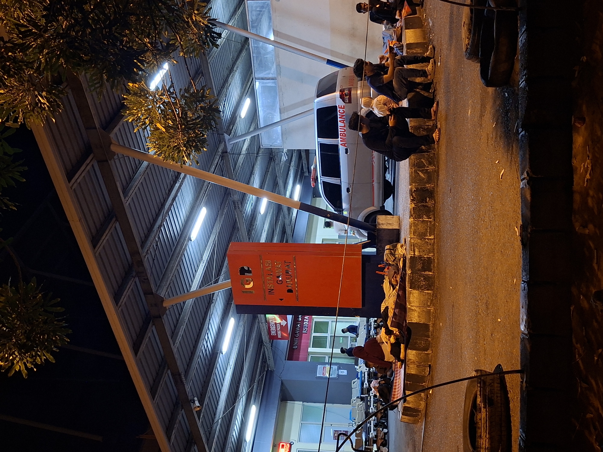 The image depicts the exterior of a hospital's emergency department at night, identified by signage as "IGD INSTALASI GAWAT DARURAT" which translates to "Emergency Installation". The location is specified as Banda Aceh, Indonesia.
The scene is illuminated by artificial lights from the hospital and overhead street lamps. Several individuals are seated and lying on the ground outside the building, some on mats and blankets, suggesting they are waiting or possibly sheltering. Among the visible subjects are at least five men, one of whom is wearing a headscarf. One man is seen holding a mobile phone.
An ambulance is parked in front of the building. In the background, multiple hospital beds are lined up under a covered walkway. The environment is paved, with decorative curbing and some debris on the ground. Overhead, trees with foliage are visible against the dark night sky. The overall impression is one of a functional, albeit somber, emergency hospital setting at night.