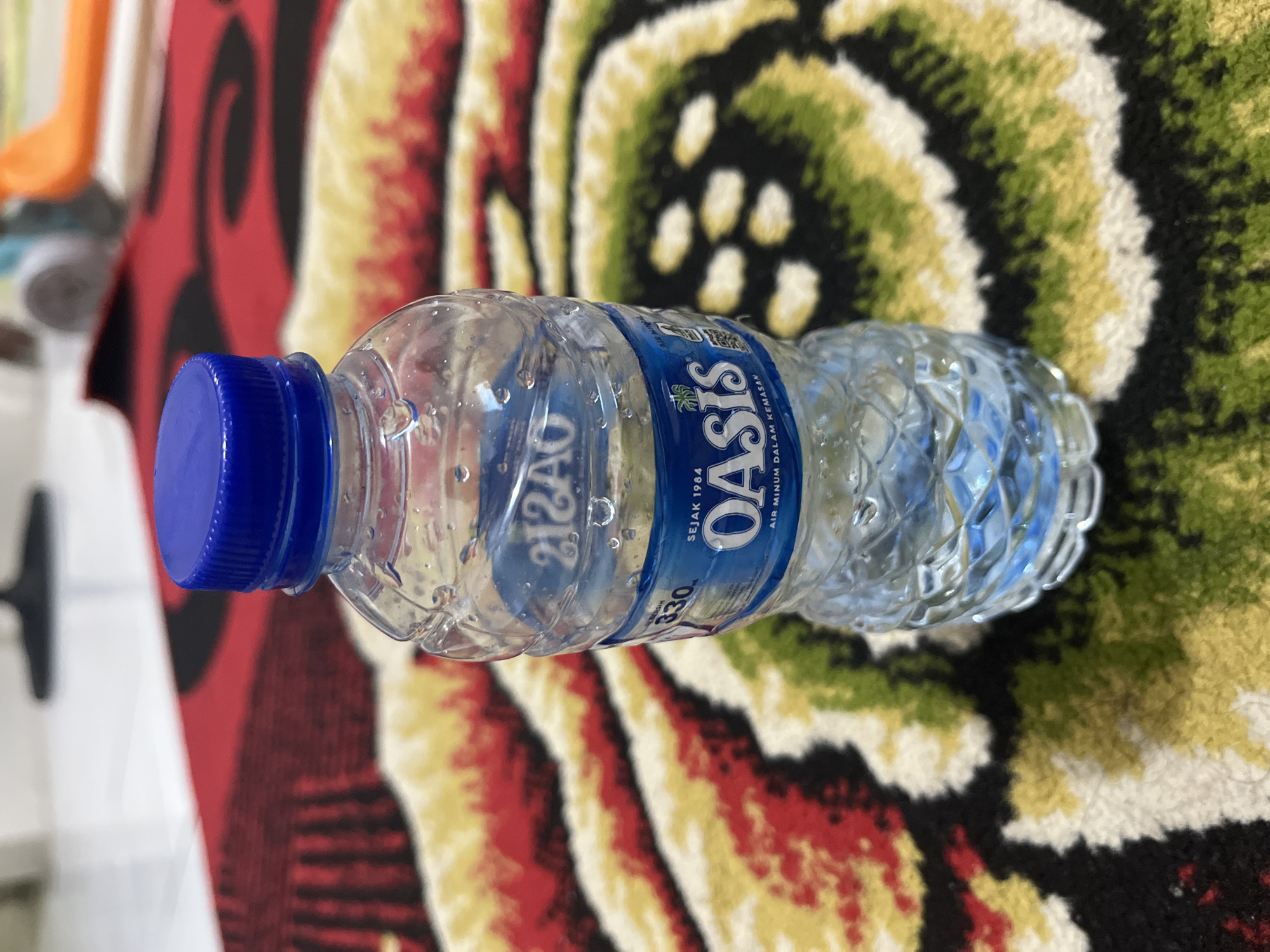 A clear plastic bottle of Oasis brand mineral water stands upright on a patterned rug. The rug features a repeating design of red, yellow, and green swirls on a black background. The bottle has a blue cap and a blue label that reads "OASIS" with "SEJAK 1984" above it and "AIR MINUM DALAM KEMASAN" below. The bottle contains clear liquid and is slightly wet, with visible water droplets on its surface. In the background, a blurred image suggests an indoor environment with furniture. The lighting is bright, indicating the photo was taken in a well-lit area. No specific details are present to determine the exact location within East Jakarta, Indonesia.
