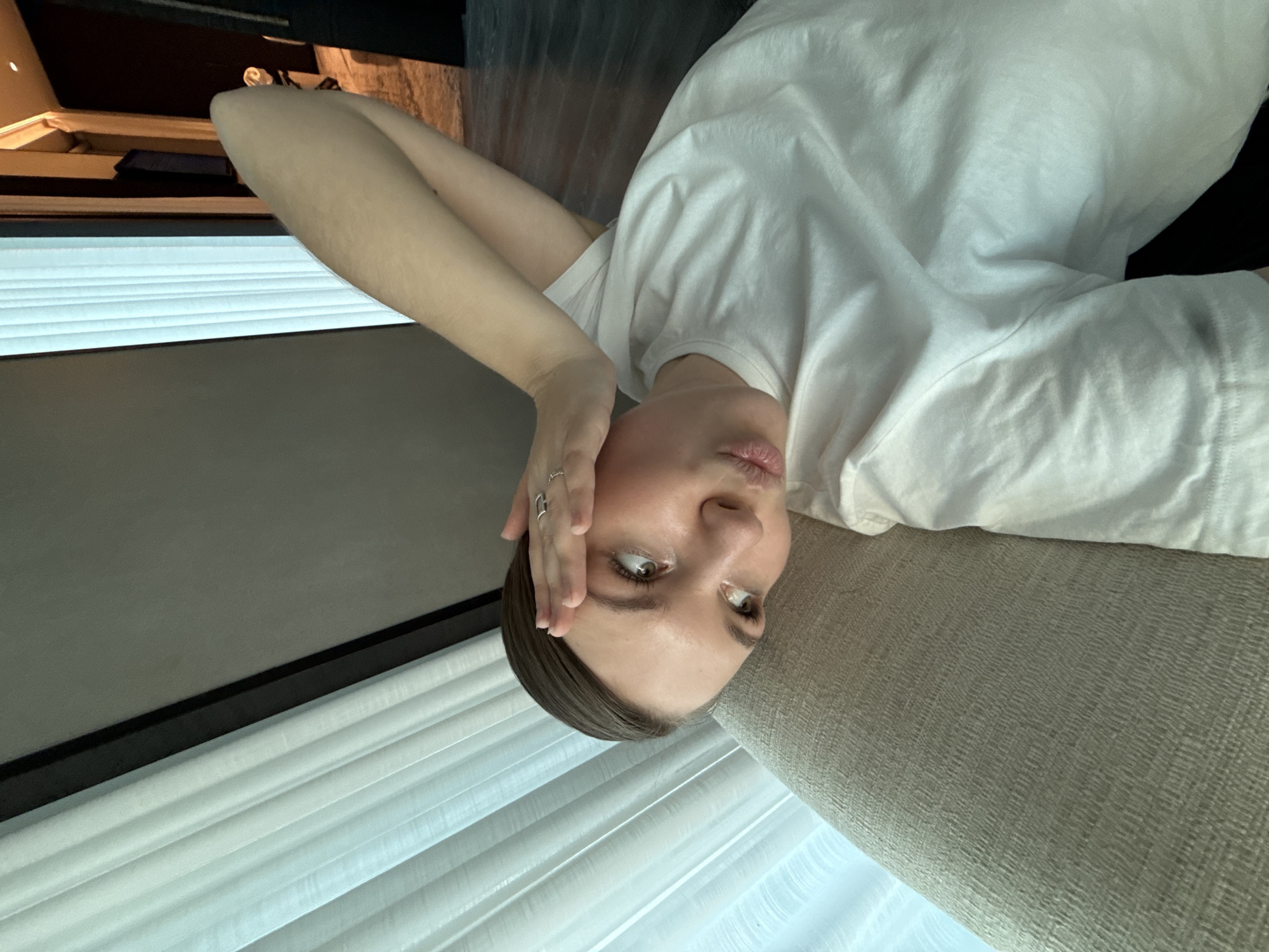 A woman with light skin, short dark hair, and green eyes is lying on a light brown textured surface, possibly a sofa or bed. She is wearing a white t-shirt and a silver ring on her hand, which is resting on her forehead. The background features a window with white blinds and a partially visible dark-colored object, possibly furniture or a TV, in the upper left corner. The orientation of the image suggests that the woman is lying down and looking at the camera. There is no indication that the location is in Vracar Urban Municipality, Serbia, based on the visual content of the image.