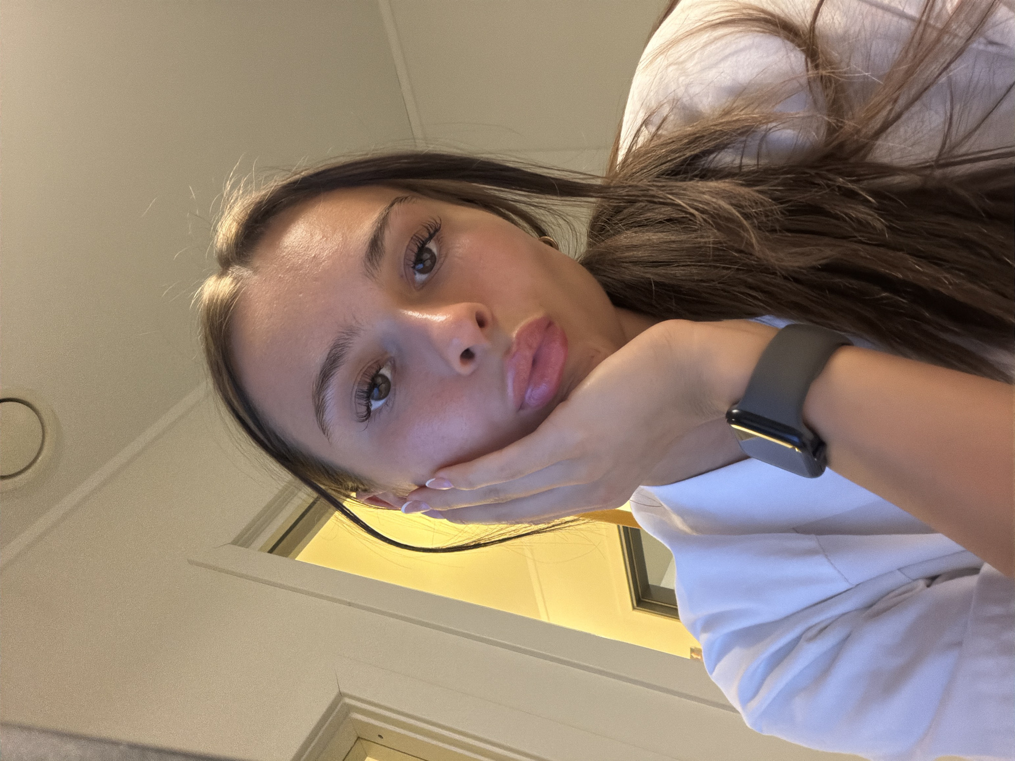 A young woman with long, brown hair and light skin is looking at the camera with her hand on her cheek and her lips pursed. She is wearing a white top and a dark smartwatch on her left wrist. The background appears to be indoors, with a cream-colored wall and a yellow-lit doorway visible. The lighting suggests it is daytime. The location is not specified in the image or its context.