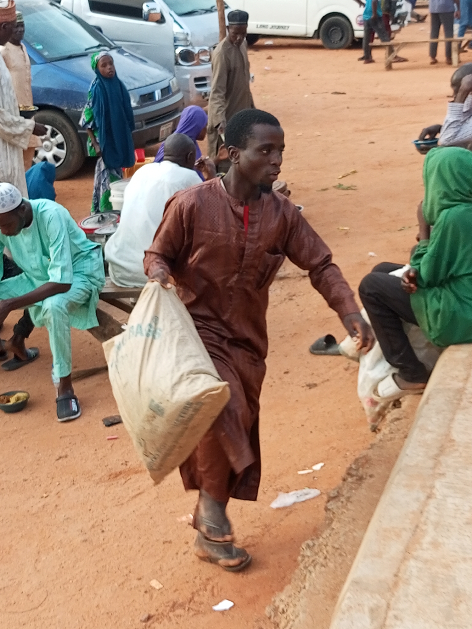 This is a medium shot, eye-level photograph taken outdoors, likely in the city of Gombe, Nigeria, featuring a group of people in a sandy, outdoor market or gathering area. The ground is composed of reddish-brown sand, with sparse patches of dry grass and small debris.
In the foreground, a man wearing a long, dark brown tunic with intricate stitching and a red accent at the collar is walking towards the right of the frame. He is carrying a large, light-colored woven bag in his right arm. His left hand is resting on the bag. He is wearing dark sandals.
To the left of the man, another person in a light blue-green tunic and pants is seated on a wooden structure. This person is wearing dark sandals. In front of them, on the ground, is a small dark bowl containing some food.
Behind the seated person, a young person wearing a blue hijab and a patterned garment is standing. To their left, a blue car is visible. Further back, other individuals are seen in various attire. A white van is parked in the background.
To the right of the main subject, a person in a bright green hijab is seated, facing away from the camera. They are wearing black pants and white sandals.
The overall atmosphere appears to be that of a busy, informal gathering, possibly a market or a public square, with people engaged in their daily activities. The lighting suggests it is daytime, with the sun casting shadows.