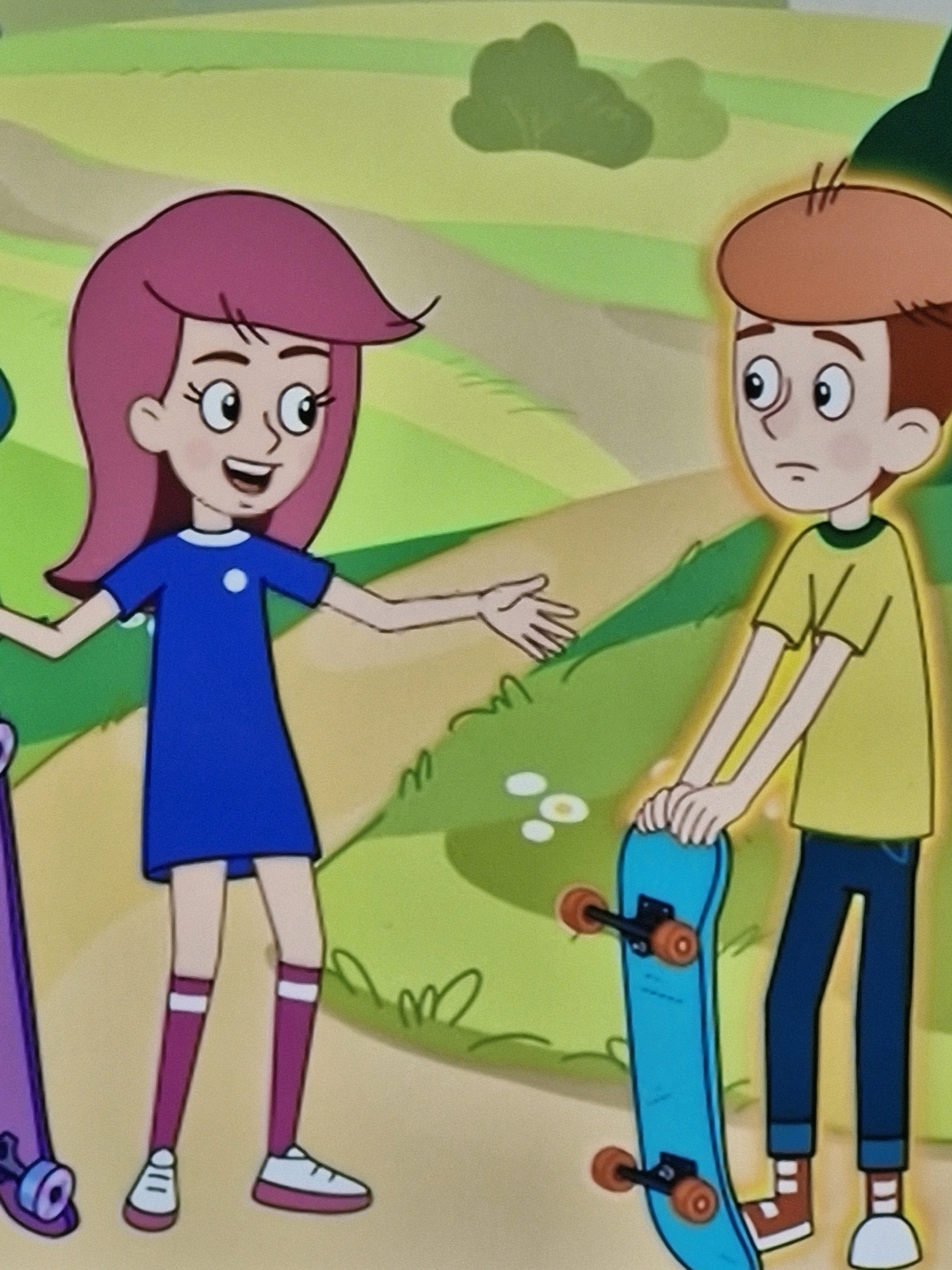 The image depicts an animated scene with two children outdoors, in what appears to be a park or a nature trail. On the left, a girl with short, vibrant pink hair, wearing a blue dress and purple knee-high socks with white stripes, stands with her arms outstretched, smiling. Next to her, on the right, a boy with reddish-brown hair, wearing a yellow t-shirt and blue jeans, holds a light blue skateboard vertically. The background consists of rolling green hills and a dirt path. The scene is brightly lit, suggesting daytime. There is no visible indication of the city of Kazan, Russia, within the image.