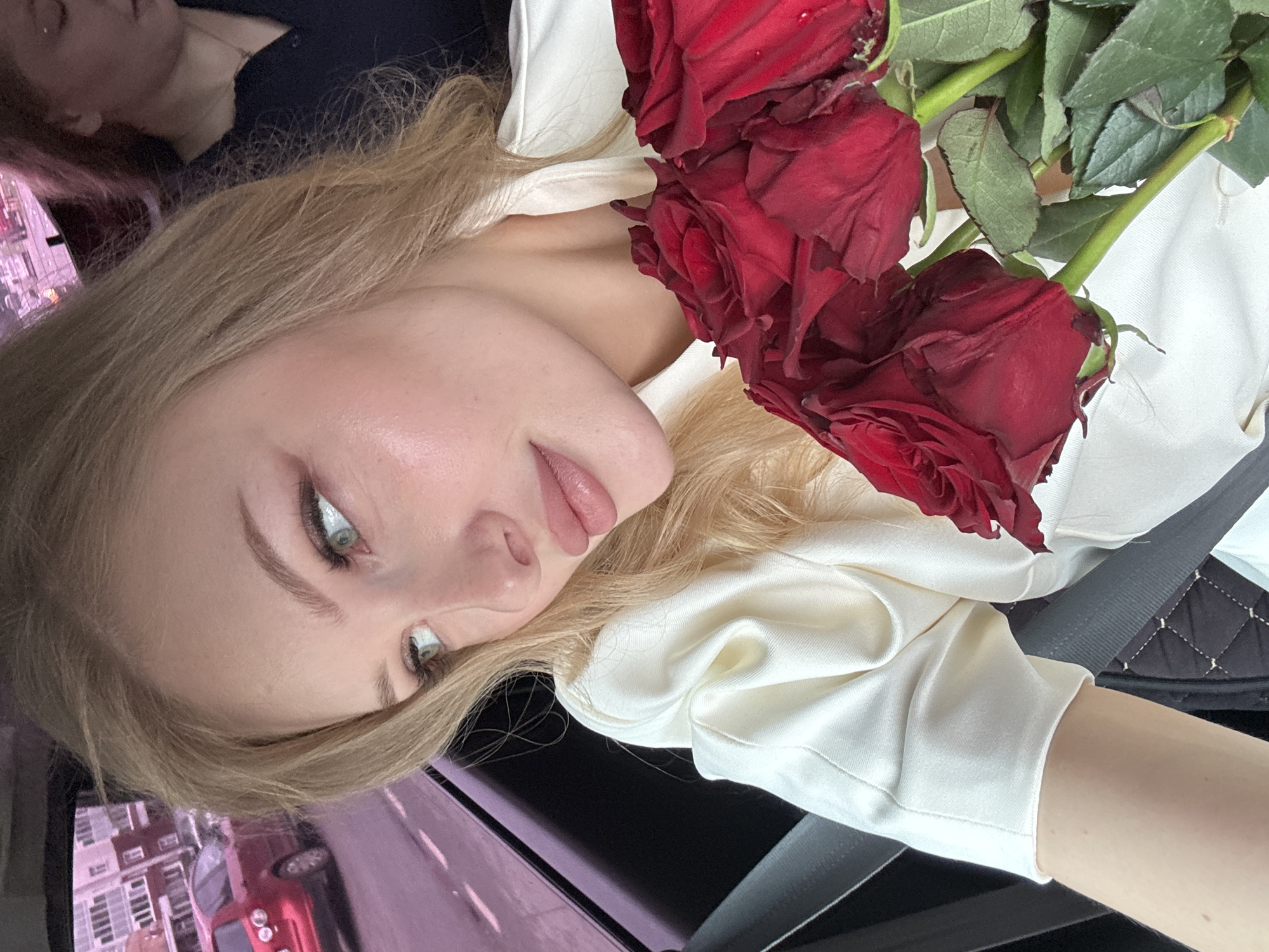 A young blonde woman is photographed from the chest up, looking out the window of a car. She is wearing a cream-colored satin blouse with puffed sleeves. She has fair skin, blue-green eyes, and light pink lipstick. Red roses are held in her right arm. In the background, through the car window, a red car is visible on a street with buildings that appear to be in a city. The location is specified as Perm, Russia. Another person, a woman with reddish-brown hair, is partially visible in the background to the right, also seated in the car. The overall lighting suggests it is daytime, possibly overcast.