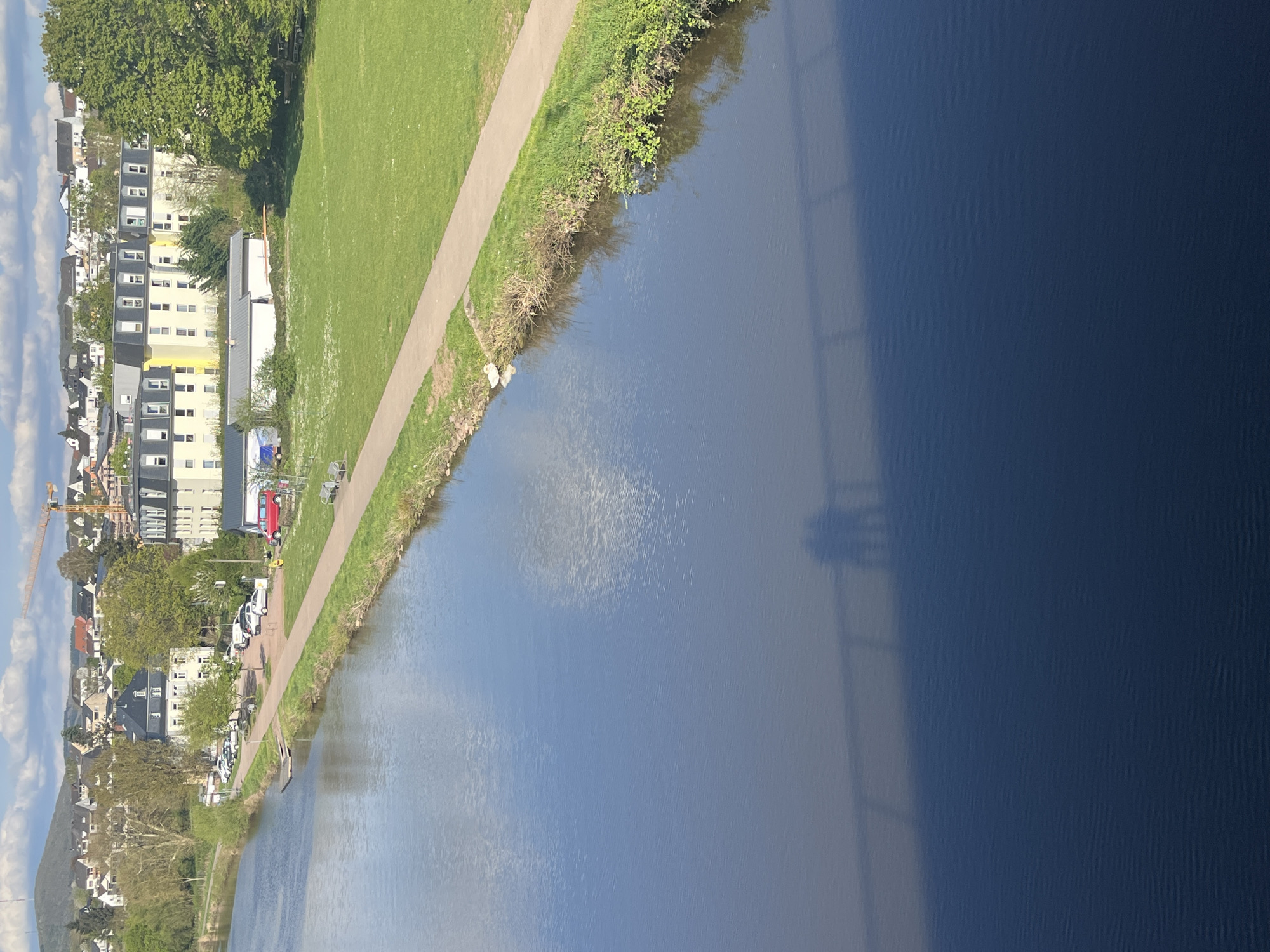 A view along the Saar River in Saarburg, Germany, shows the river on the right of the frame, reflecting the sky. The far bank is visible on the left, with buildings, trees, and a path running along the river. The path is adjacent to a grassy area, and cars are parked along the path on the far left. A construction crane is visible in the distance. The right side of the river is in shadow, obscuring the details. The image is rotated ninety degrees counterclockwise.