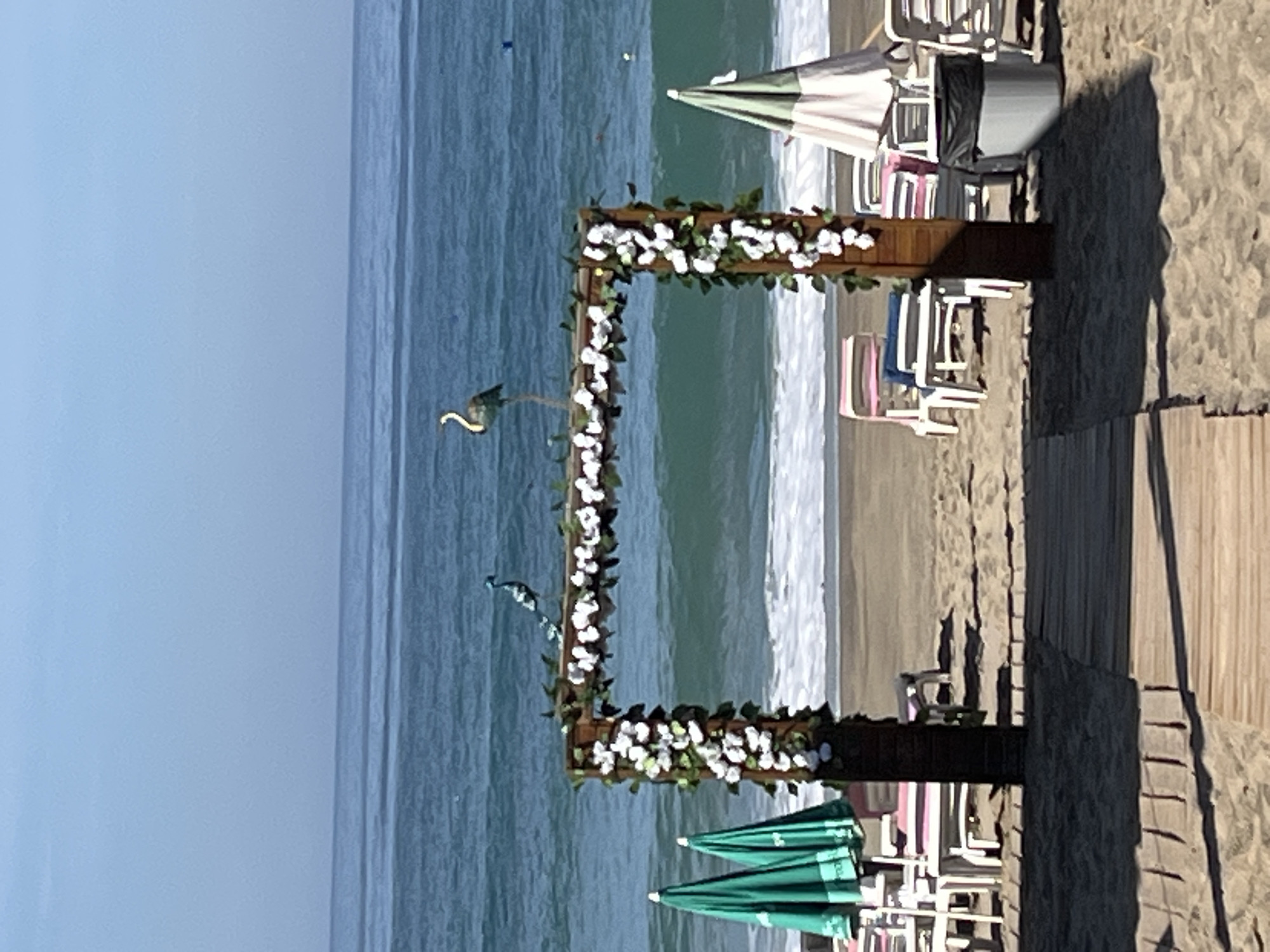 The image shows a beach wedding setup at a beach in Alanya, Turkey. A wooden arch decorated with white flowers and greenery stands on the sand. There are two decorative bird figurines attached to the arch. Beach chairs and umbrellas are arranged on the sand, and a wooden walkway leads to the arch. The sea is in the background, with a clear blue sky above. The photograph appears to have been taken during daylight hours.