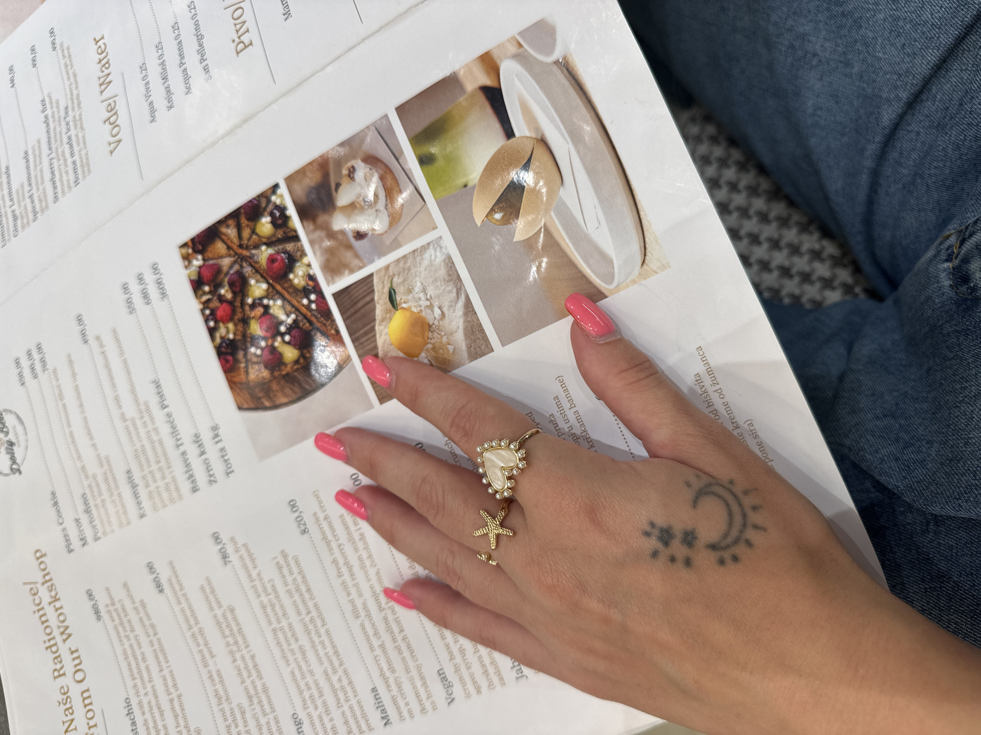 The image shows a close-up of a menu from "Naše Radionice / From Our Workshop," a business located in Belgrade, Serbia. A person's hand with bright pink nail polish and gold rings is positioned over the menu. The menu lists various food and beverage items with prices in Serbian Dinars. The background shows a grey and white patterned fabric, likely upholstery, and a person's blue denim-clad leg. The overall scene suggests the person is seated in a cafe or restaurant in Belgrade, perusing the menu.